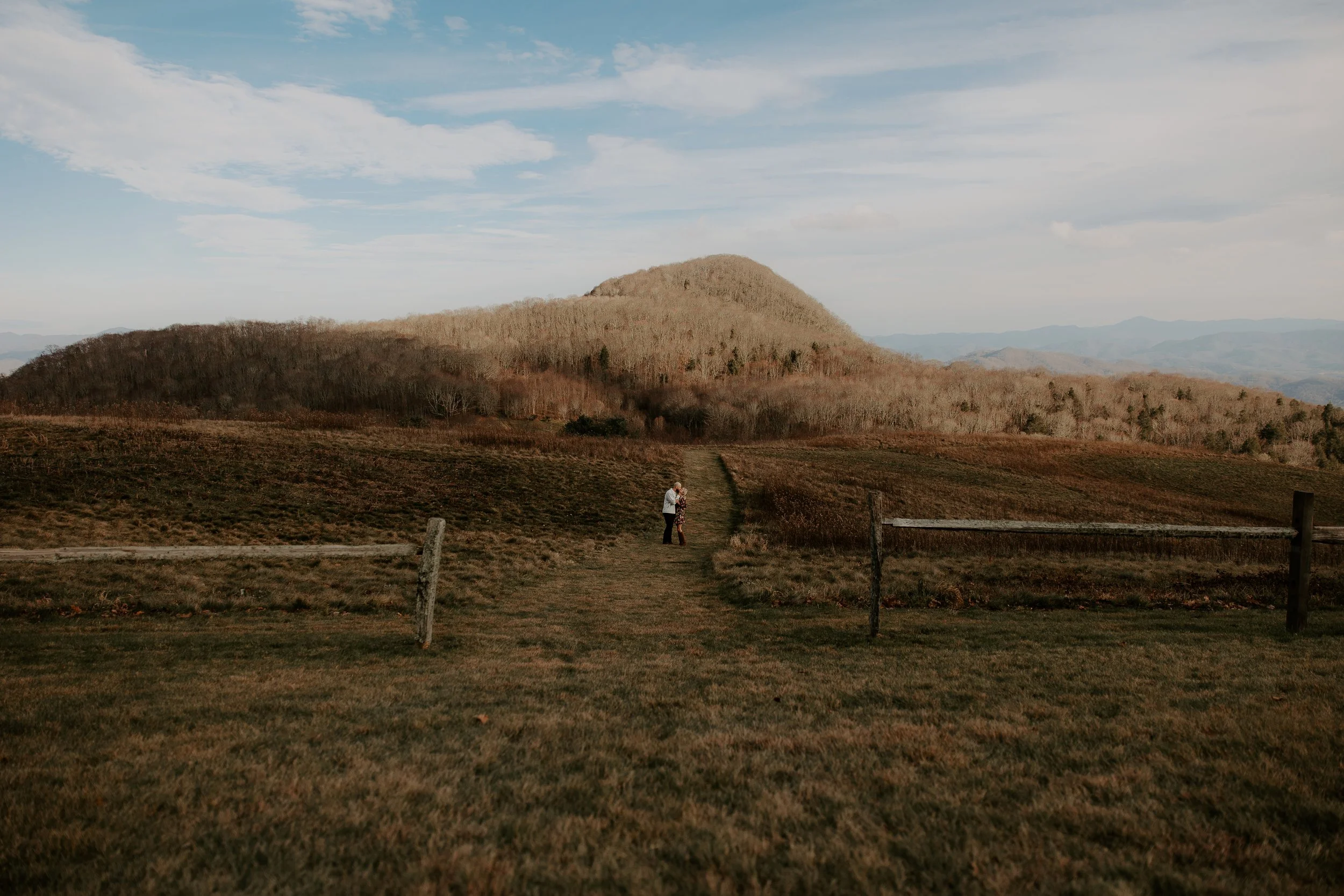 Wide landscape shot of Purchase Knob with couple barely visible on a grass path leading toward the mountain summit, split rail fence in the foreground