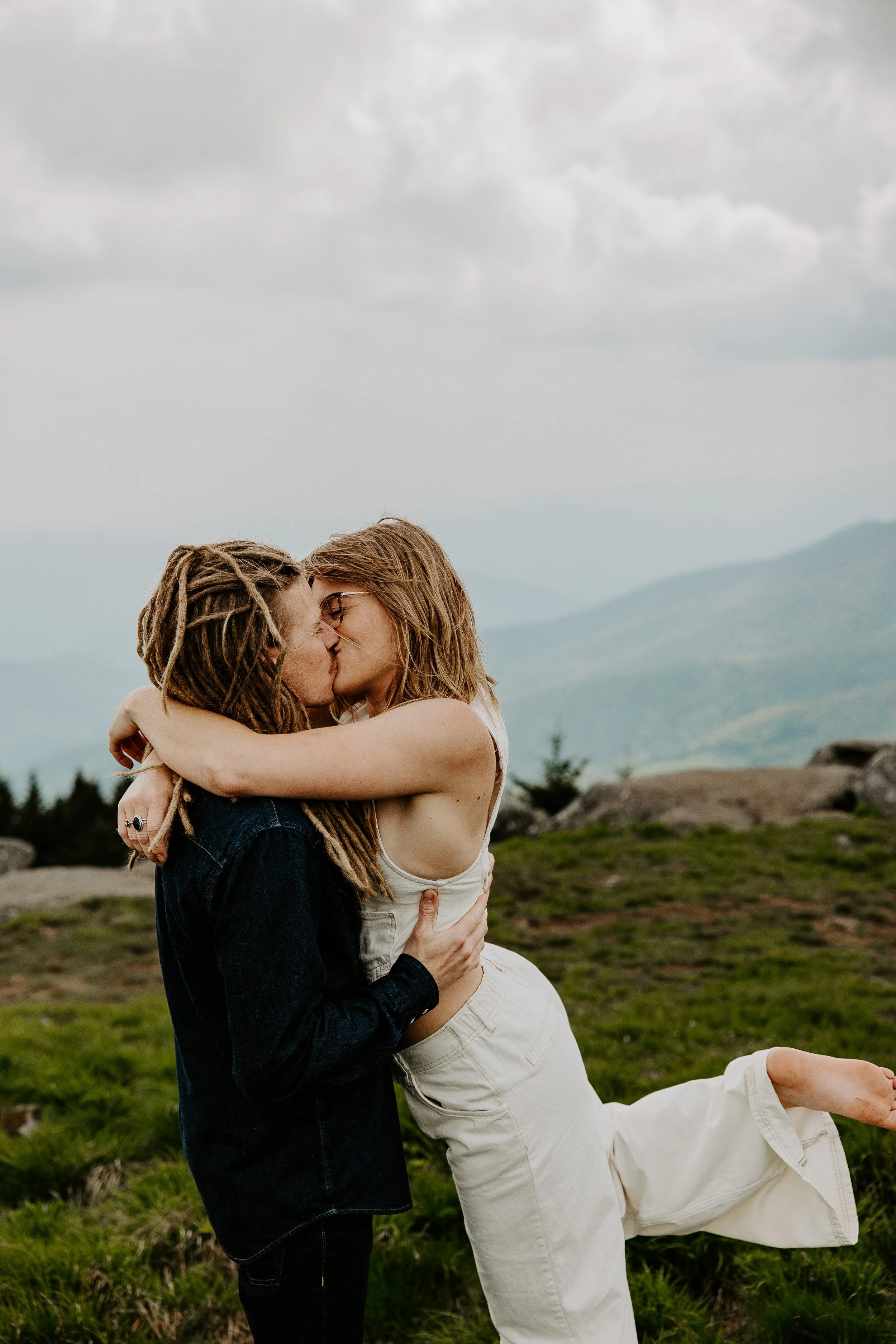 Couple kissing on a mossy green mountain bald with one partner lifting a leg, Appalachian ridgelines visible in the soft background