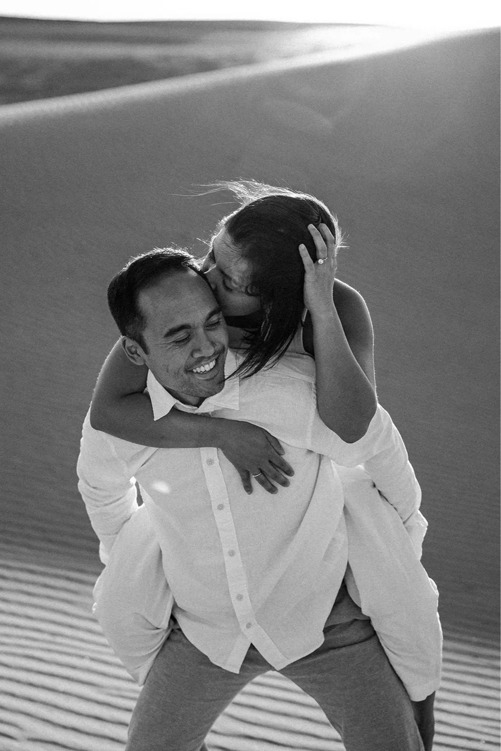 Black and white photo of a couple sharing a piggyback moment on soft sand dunes