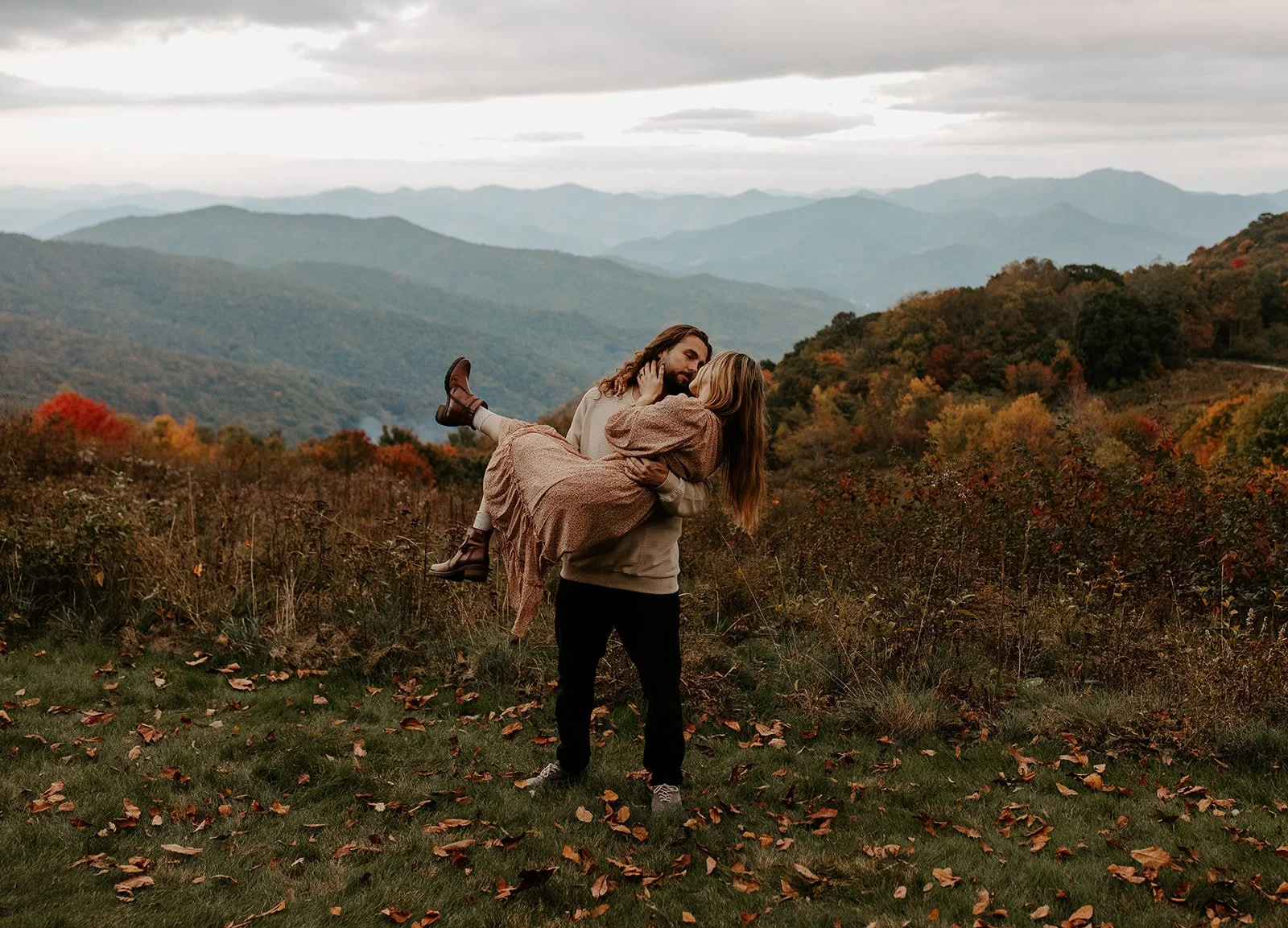 Couple embracing on an open grassy mountain bald in the Appalachians during fall, with layered ridges and vibrant autumn color spreading across the valley below