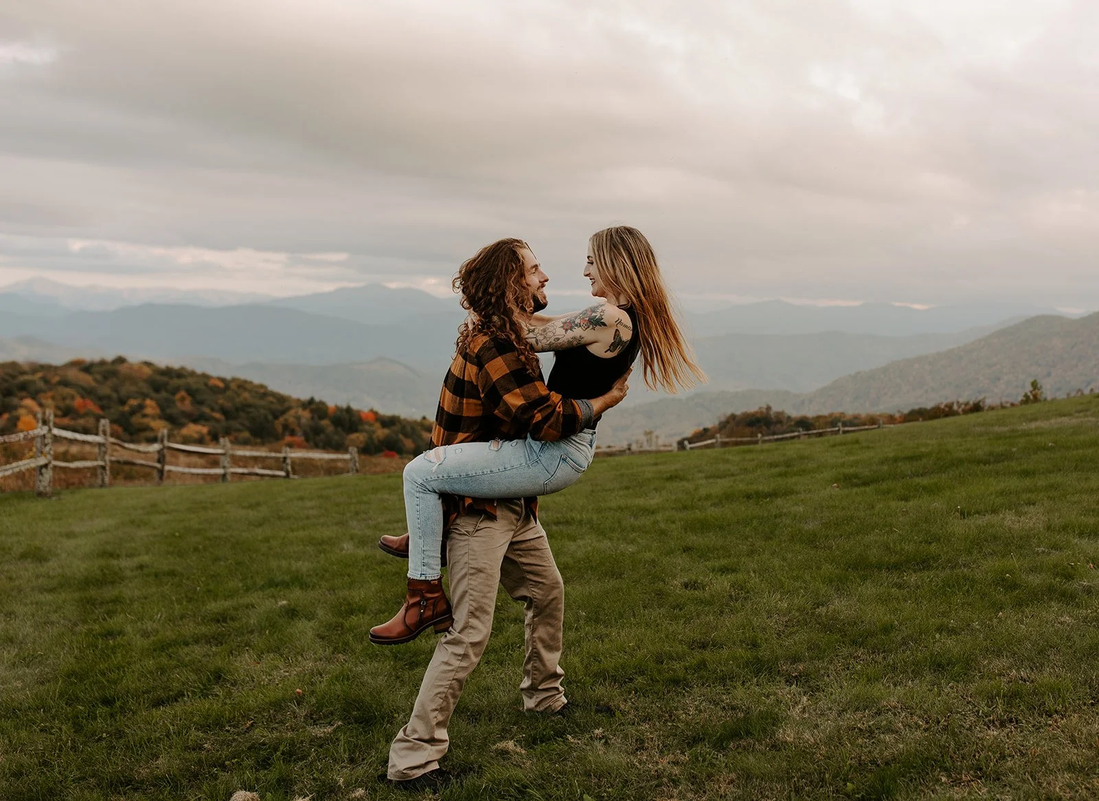 Couple laughing as one partner lifts the other in a green meadow with fall foliage and Smoky Mountain ridgelines in the background