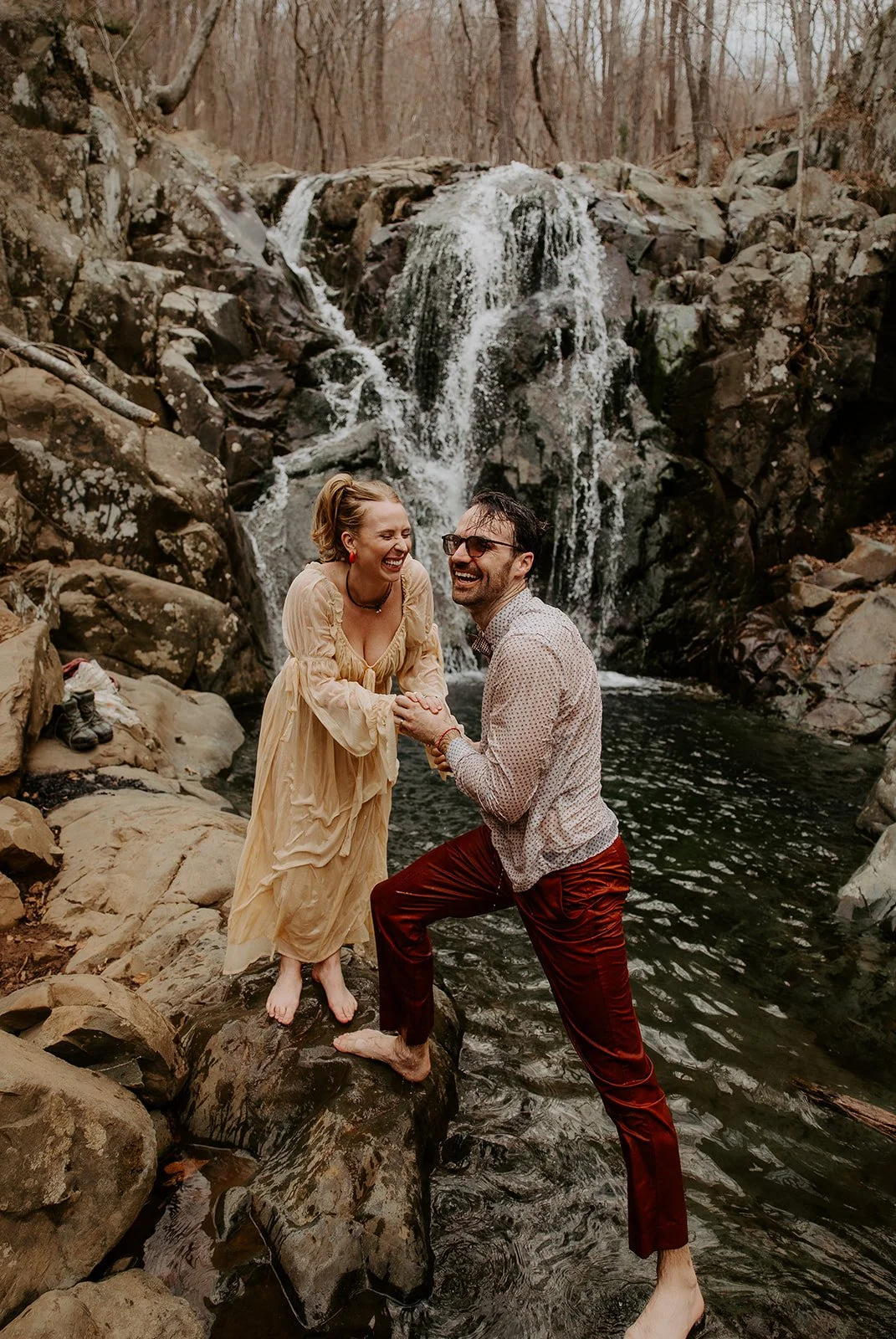 Couple laughing barefoot on rocks in front of Rose River Falls waterfall during an intimate adventure elopement