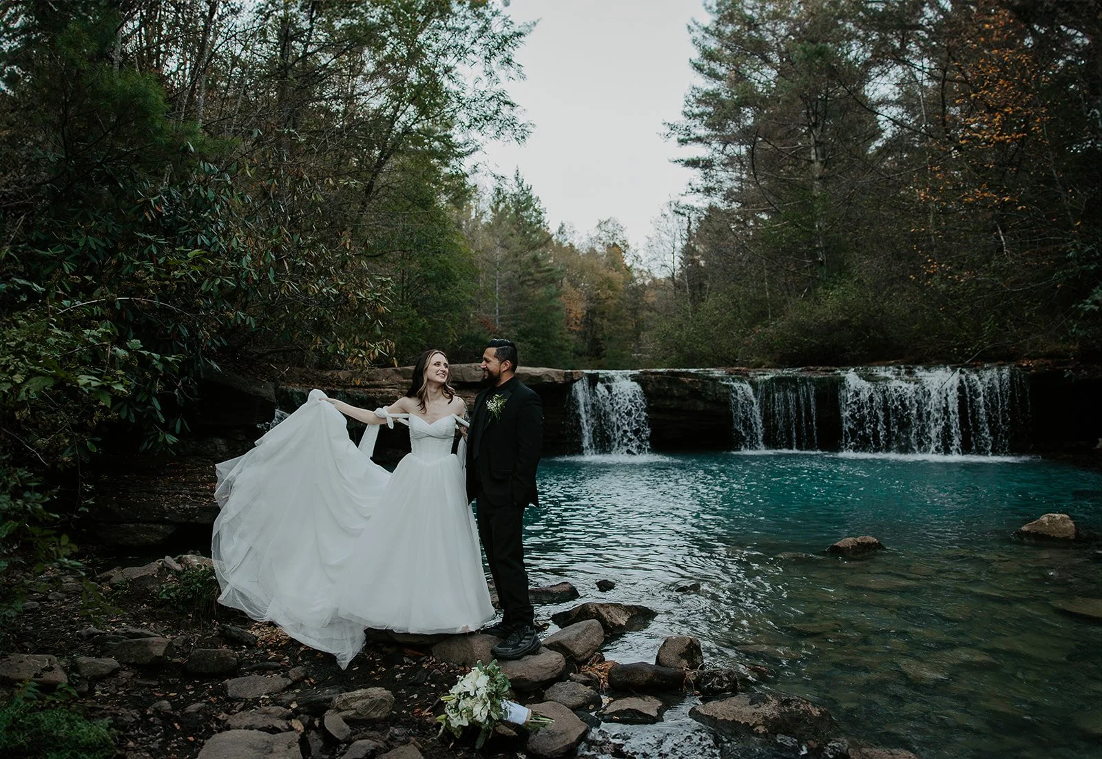 Couple eloping at a West Virginia waterfall, bride in a white ball gown and gloves standing beside a wide cascading waterfall surrounded by autumn forest