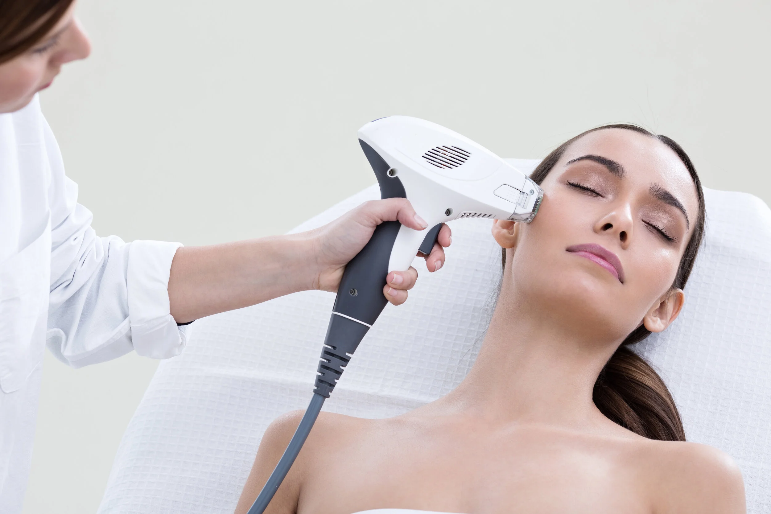 A woman receiving a skincare treatment with a handheld device on her face, lying on a white treatment bed in a clinic setting.