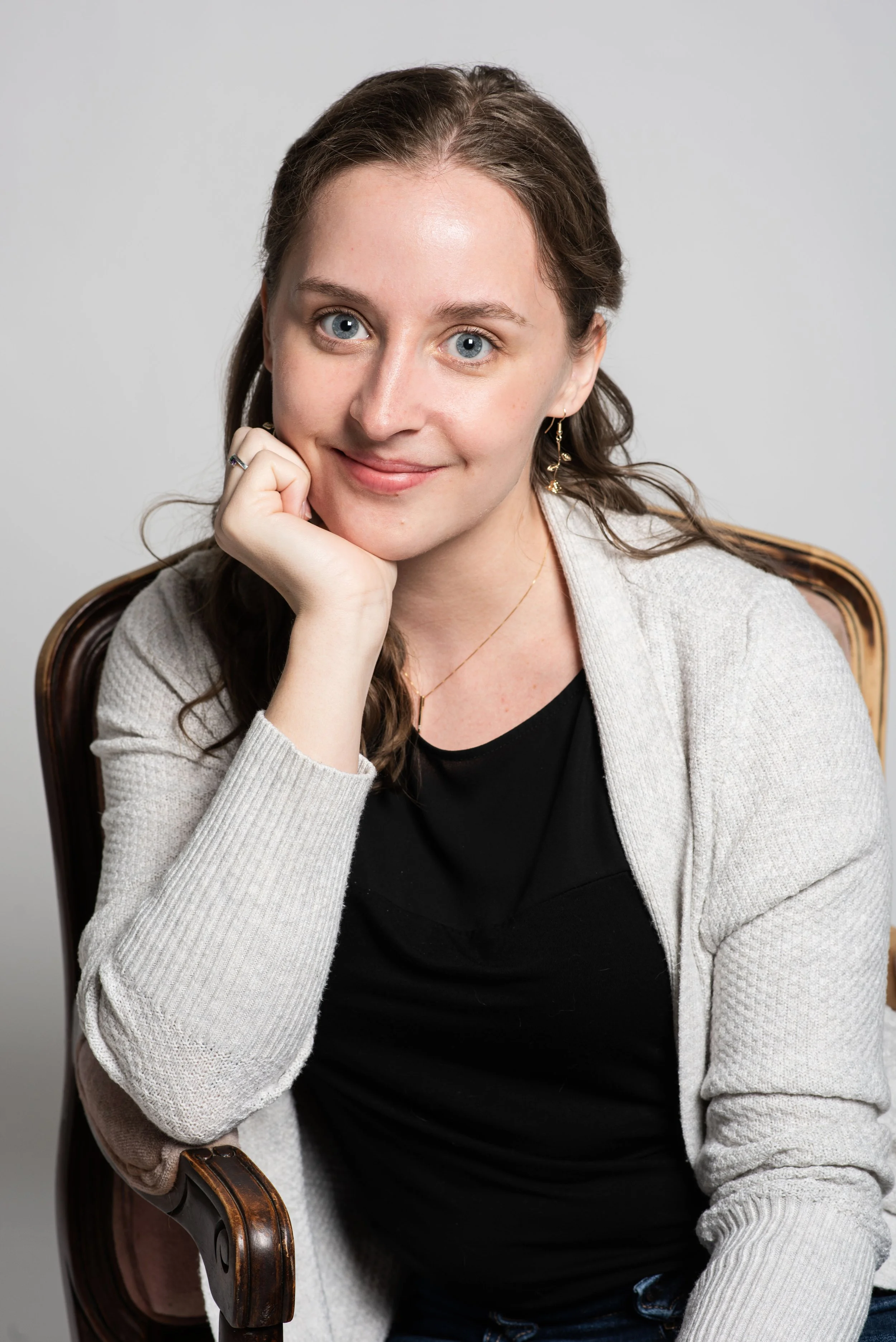Rachel, a white, feminine person is seated in a chair. Their head is resting on their hand in a therapeutic posture. They are wearing a black shirt, and a white cardigan sweater.