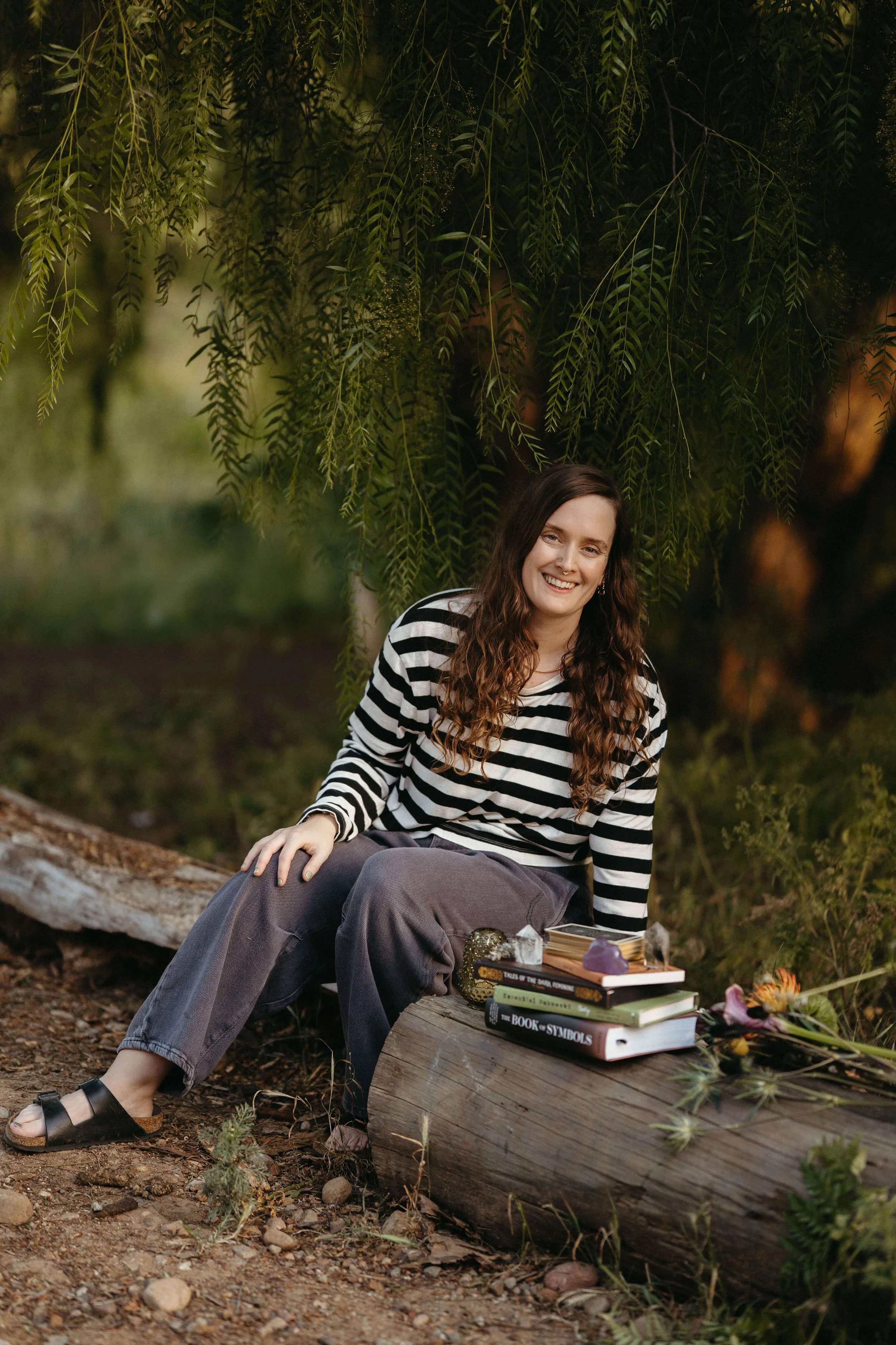 Lea M. Roman, licensed therapist and founder of Amethyst Holistic Counseling, sitting on a log surrounded by books, crystals, and a tarot deck
