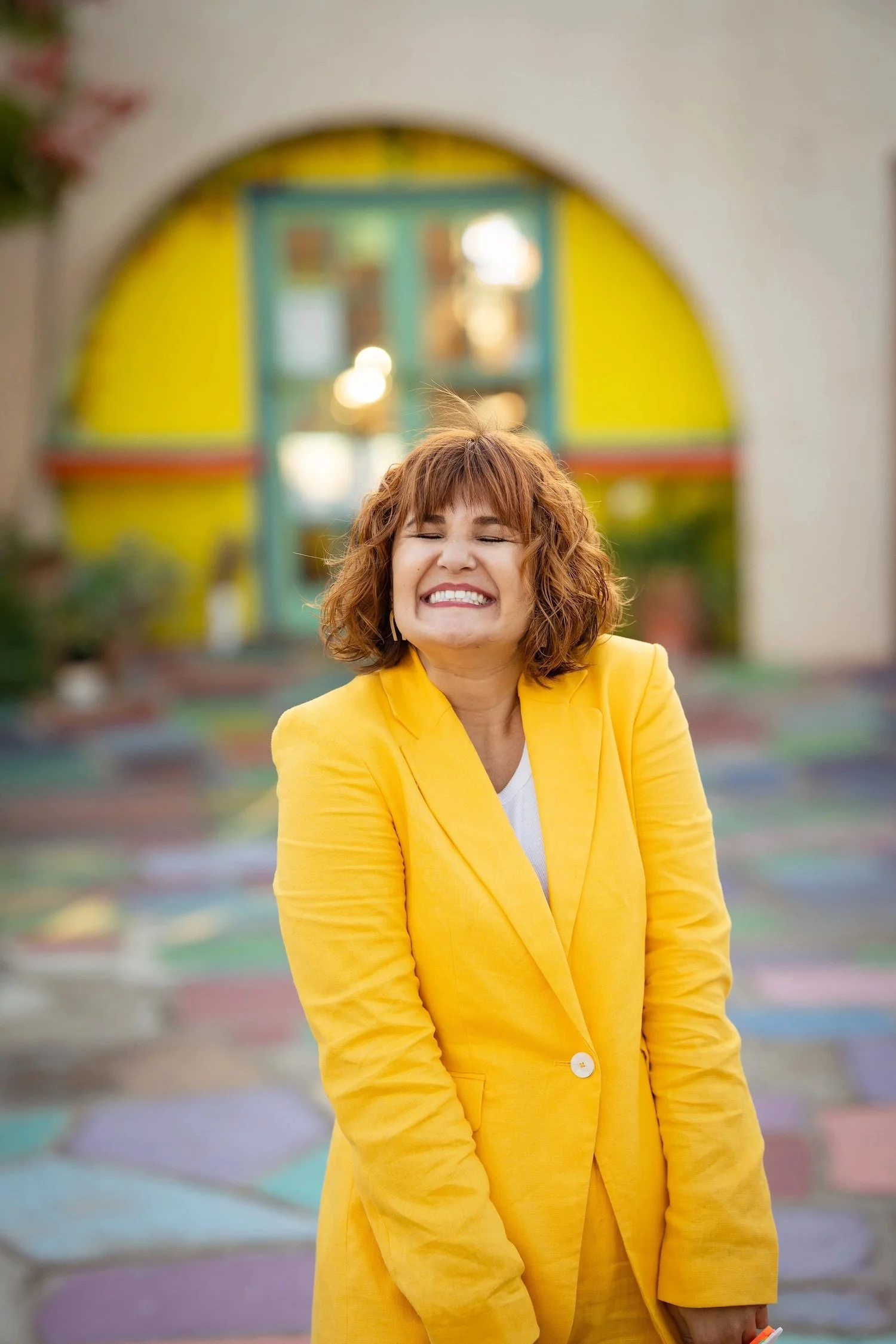Woman laughing with eyes closed in a bright yellow suit in front of a yellow arched doorway and colorful painted tiles