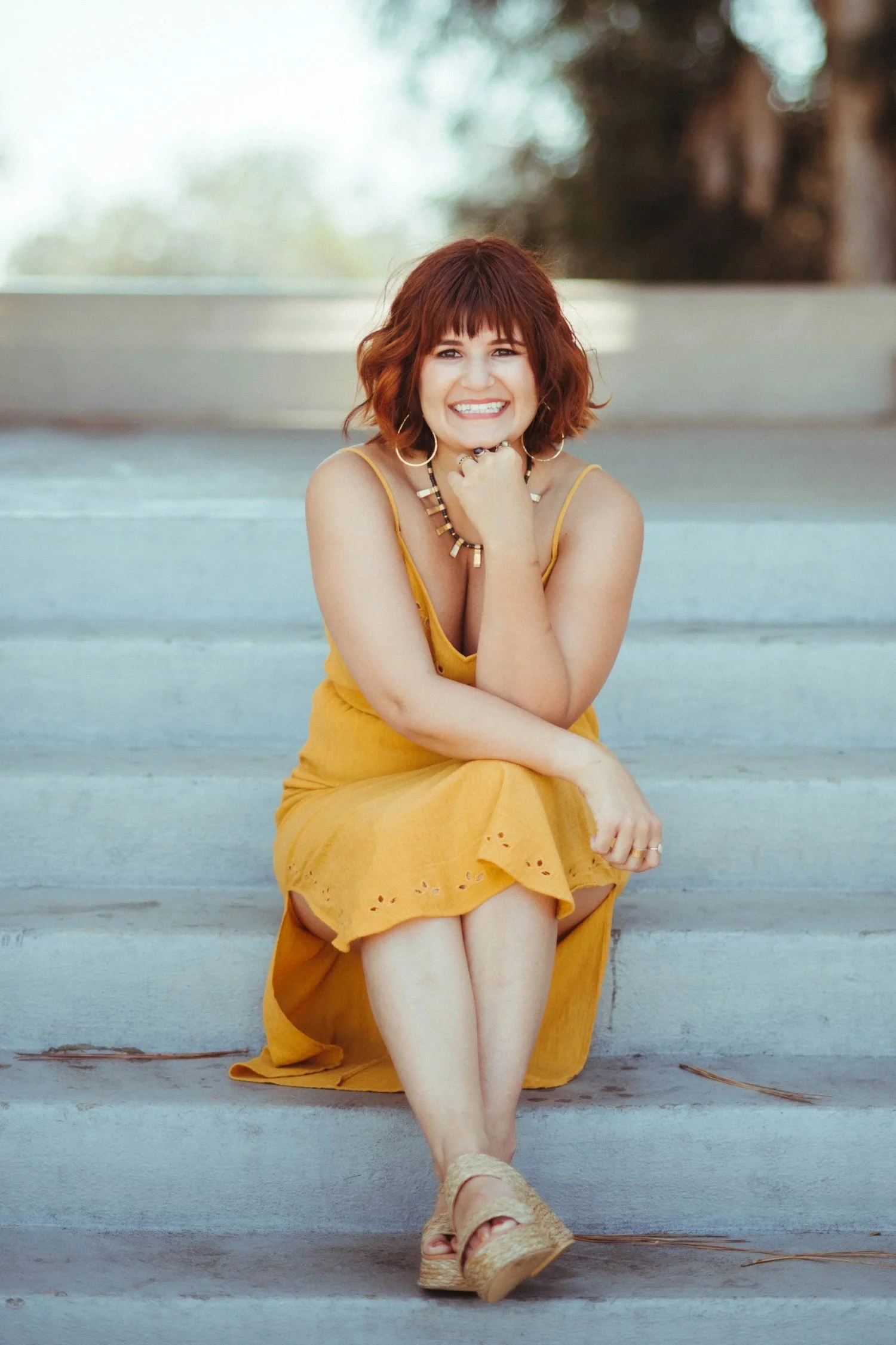Woman smiling on concrete steps in a mustard sundress with gold hoop earrings
