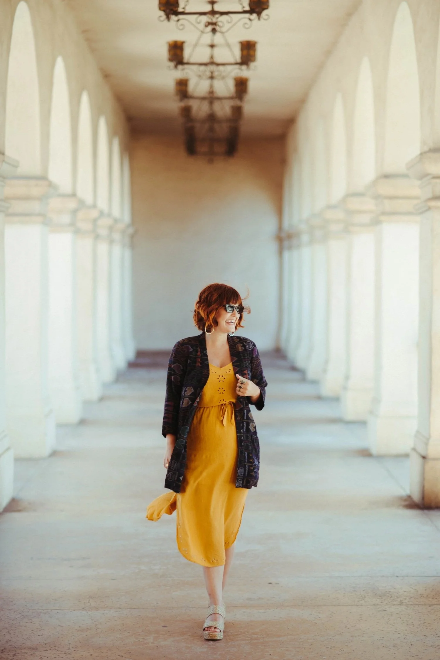 Woman smiling as she walks through a white arched colonnade in a mustard dress and patterned jacket