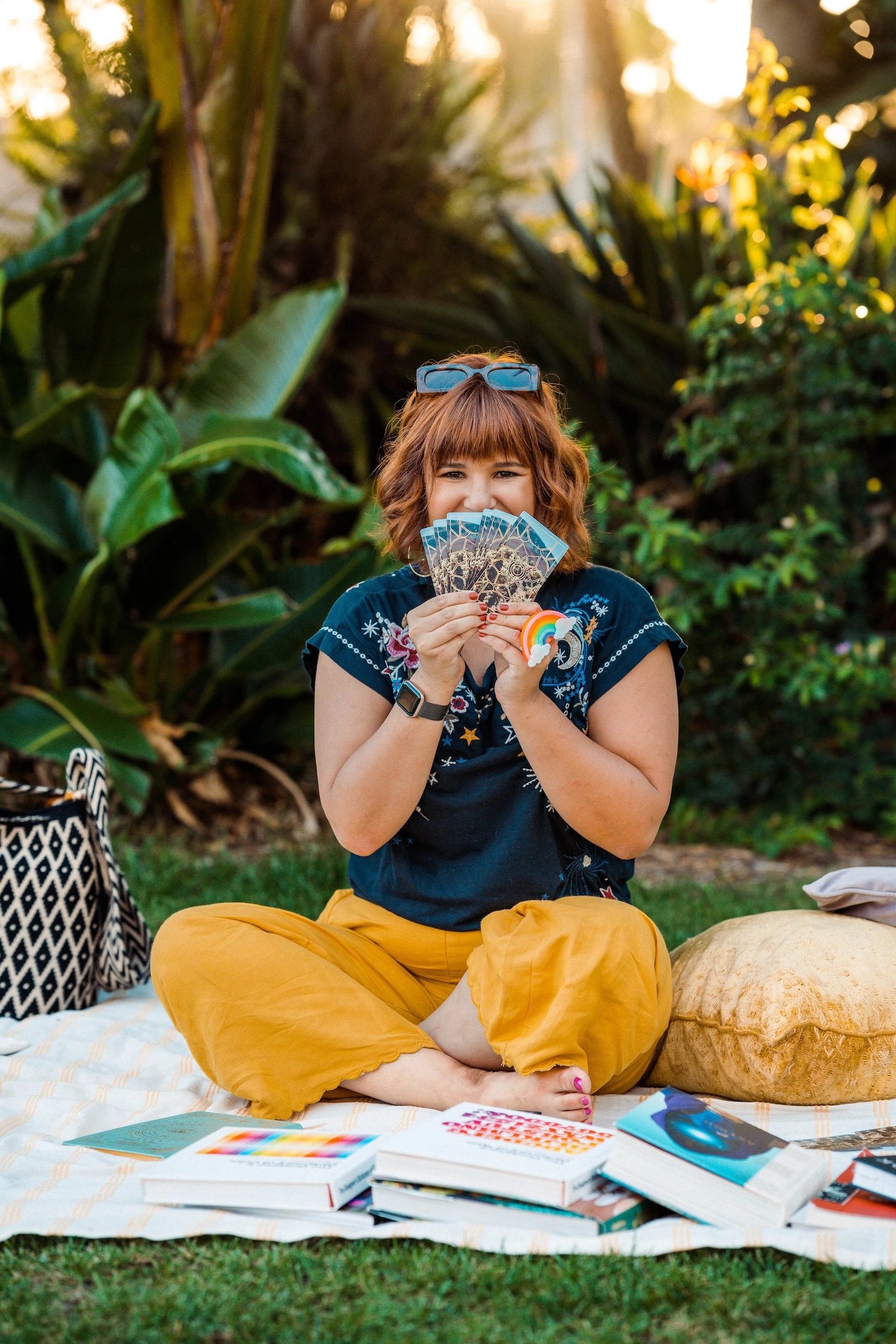 Woman peeking over a fan of tarot cards on a picnic blanket surrounded by design books in a sunlit garden