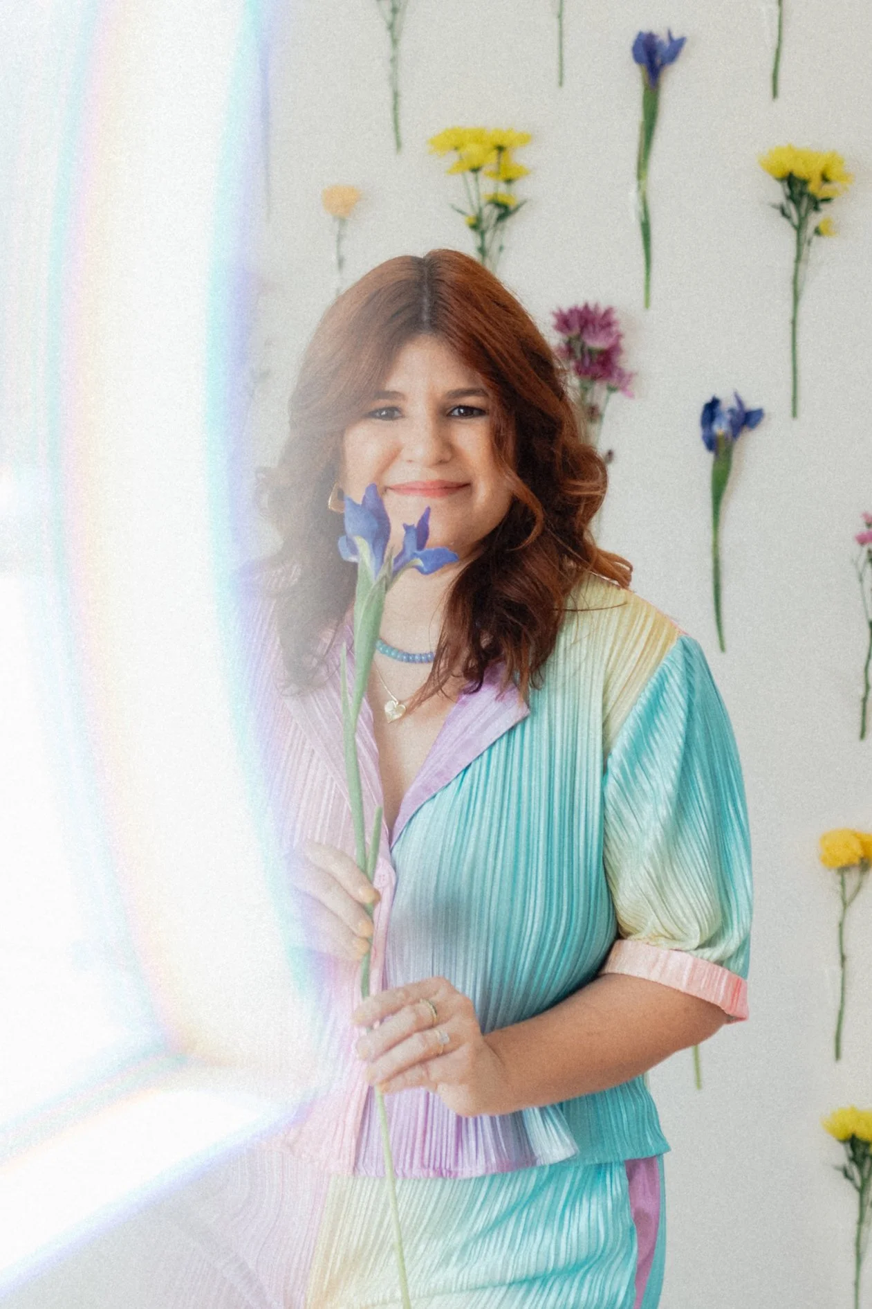 Alex McGinness, founder of Arcoíris Design Studio, smiling while holding a blue iris in a pastel rainbow outfit, with a rainbow light flare and a wall of fresh flowers behind her