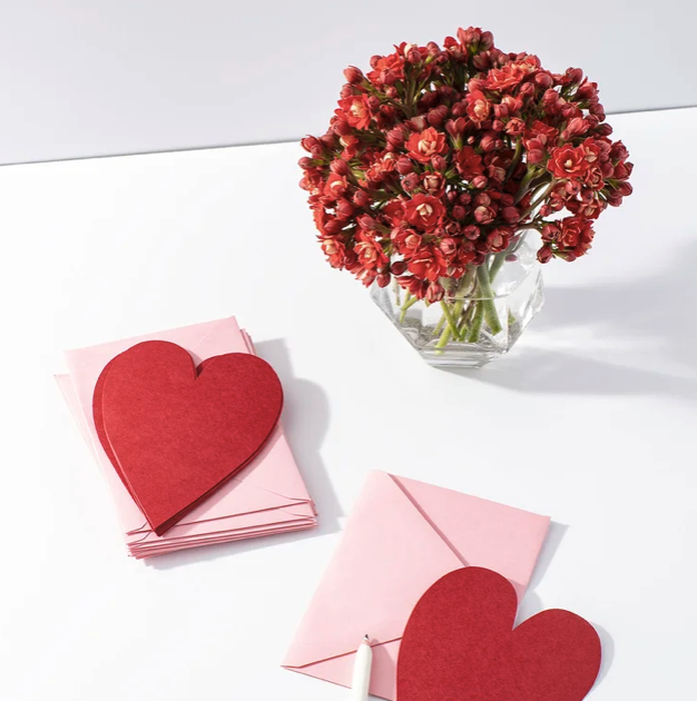 A bouquet of pink and red flowers in a glass vase, along with pink envelopes and red heart-shaped cards on a white surface.