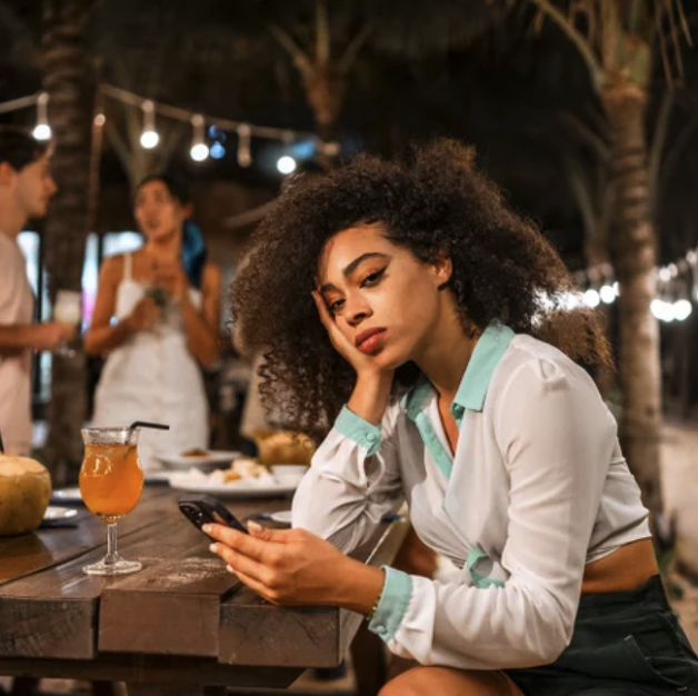 A woman with curly hair is sitting at an outdoor table and looking at her phone with a bored or annoyed expression. In the background, there are two women and string lights at night.