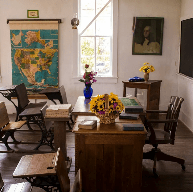 A vintage-style classroom with wooden desks and chairs, a window letting in natural light, a large map of the United States on the wall, and various flowers in vases on the desks and side table, suggesting a historical or educational setting.