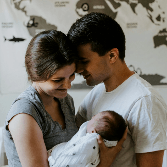 A couple holding a newborn baby, smiling and looking at each other indoors with a world map on the wall behind them.
