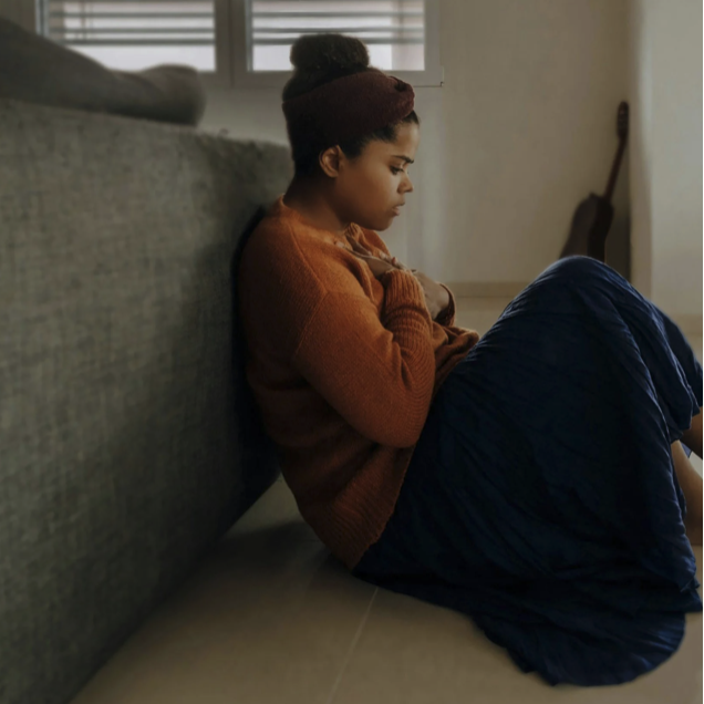 A young woman with dark hair, wearing a maroon beanie and rust-colored sweater, sits on the floor leaning against a sofa with her knees pulled to her chest and her arms crossed, appearing contemplative or sad.