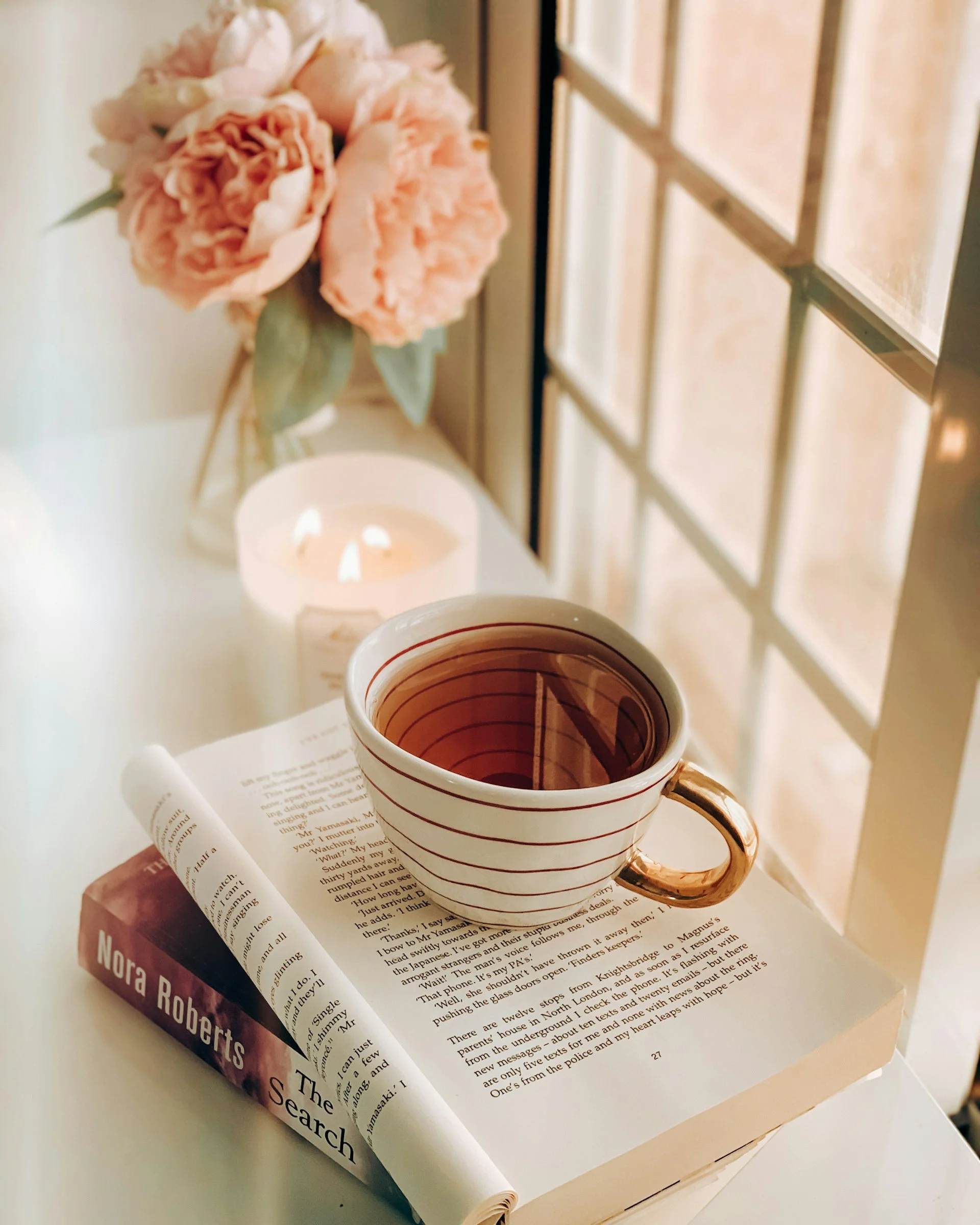 A cup of tea placed on an open book with reading glasses nearby, with a pink peony flower in the background and a window also visible.