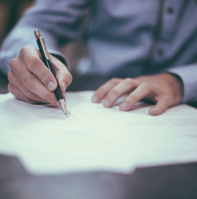 Person writing on paper with a pen, wearing a button-up shirt.