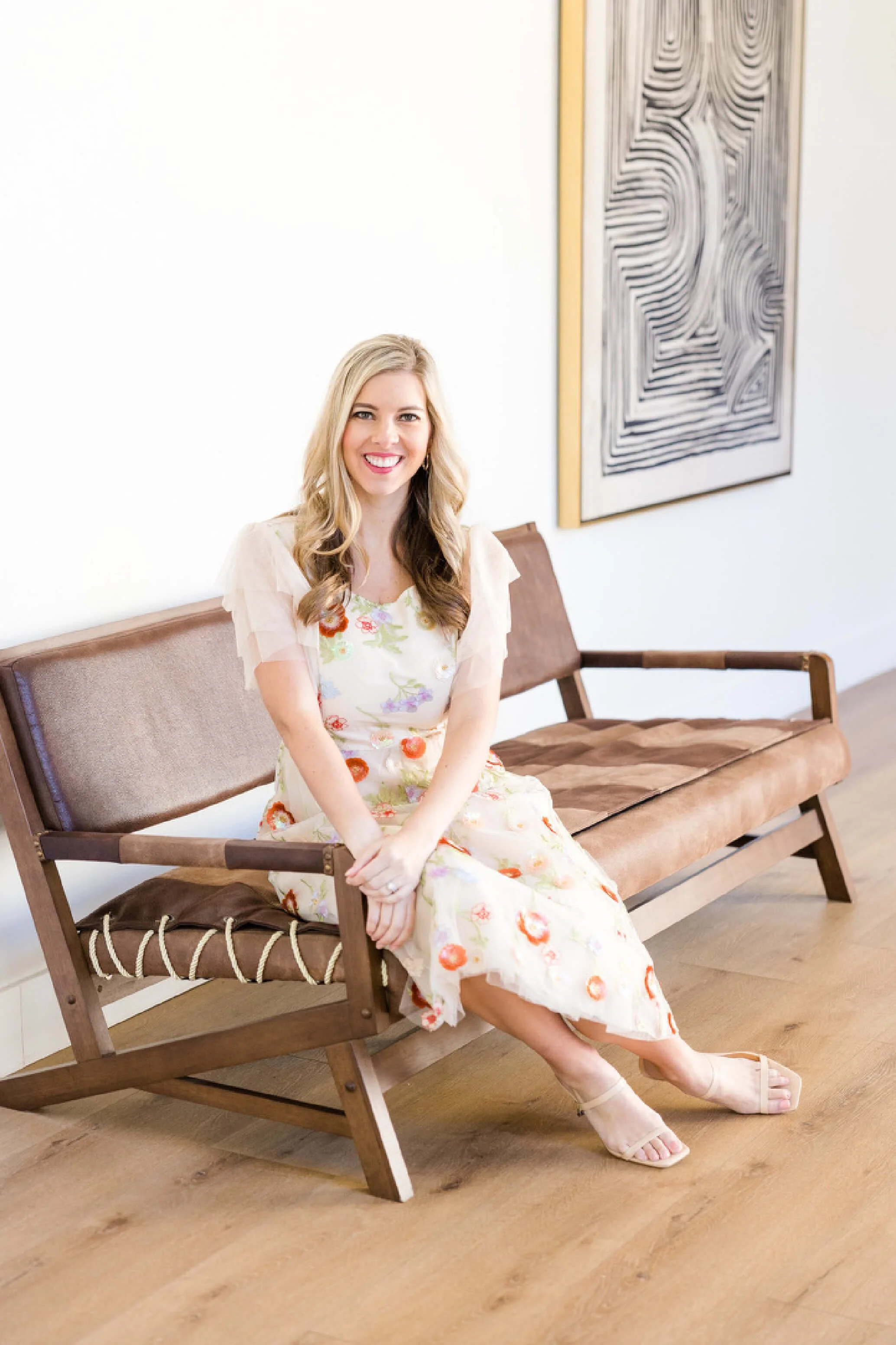 A woman with long blonde hair sitting on a wooden bench with a brown cushion, smiling, wearing a floral dress, and beige sandals. There is modern abstract artwork on the white wall behind her.