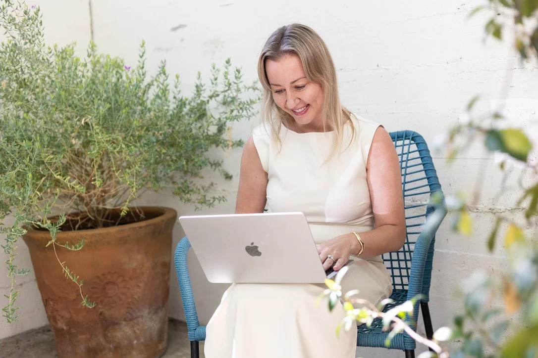 A woman sitting outside on a blue chair, using a silver MacBook laptop, surrounded by green plants.