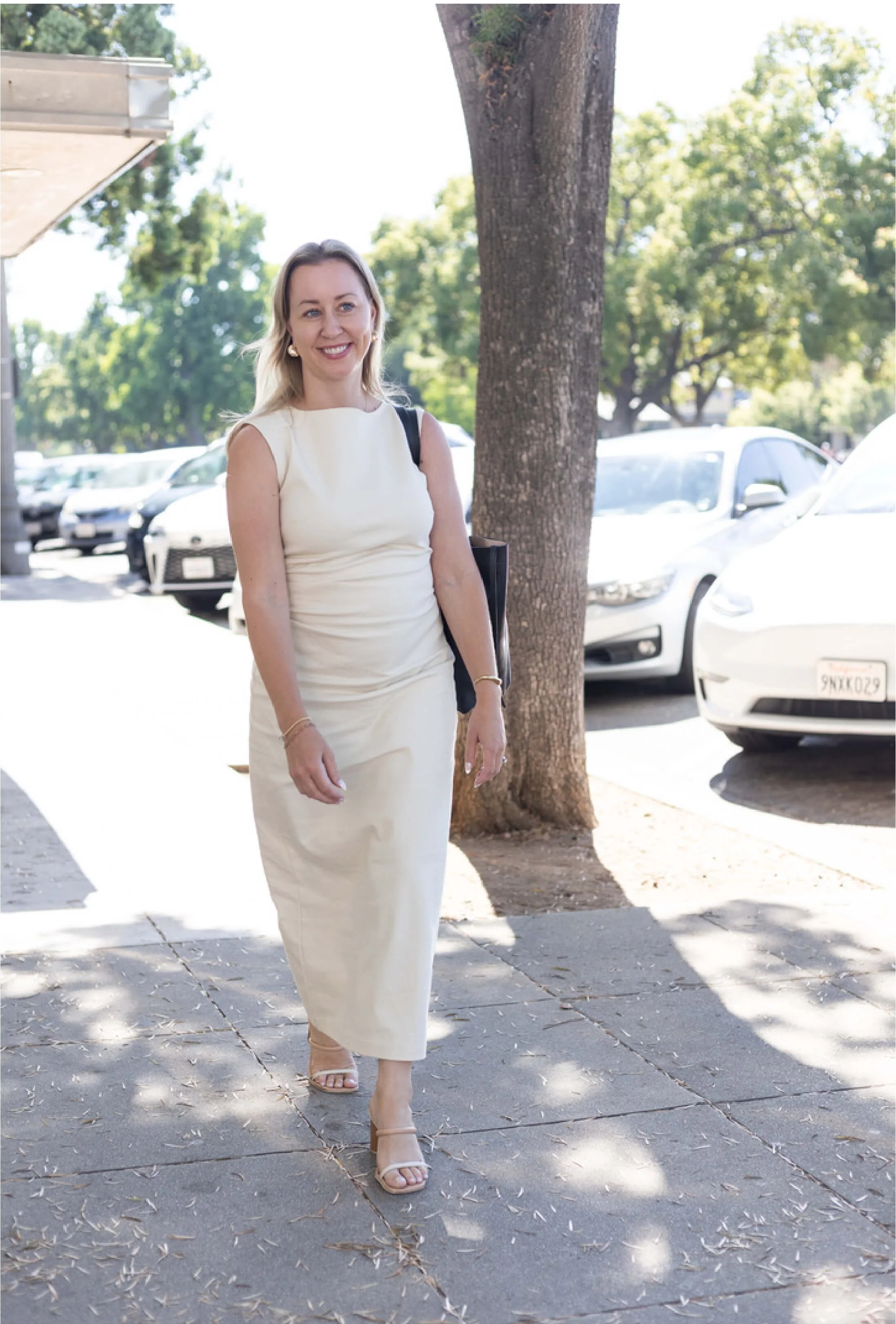 A woman in a sleeveless white dress walking on a sidewalk shaded by trees in a parking lot with several cars.