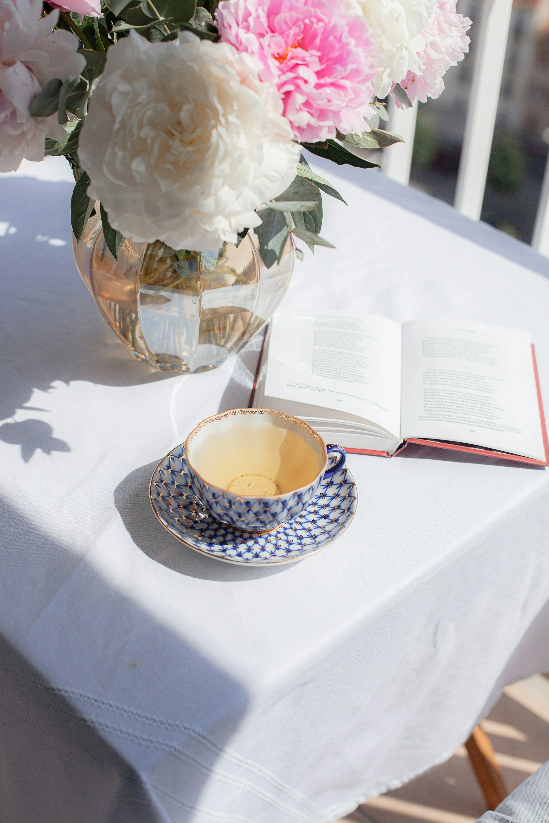 A table with a vase of pink and white peonies, an open book, a teacup with a saucer, and a teabag inside the cup, all on a white tablecloth near a window with sunlight.