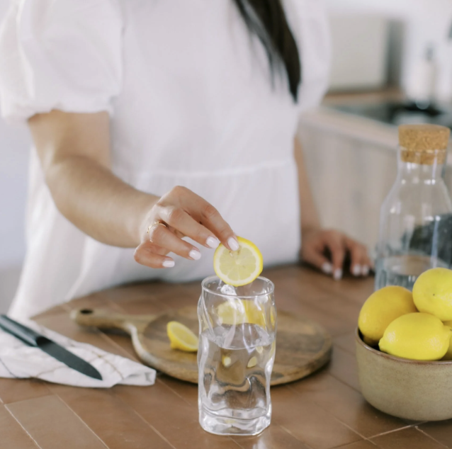 A woman is placing a slice of lemon into a glass of water on a wooden kitchen countertop with a bowl of lemons and a water bottle nearby.