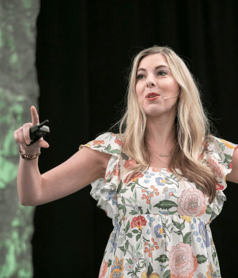 A woman with long blond hair, wearing a floral dress, giving a presentation on stage with a microphone headset.