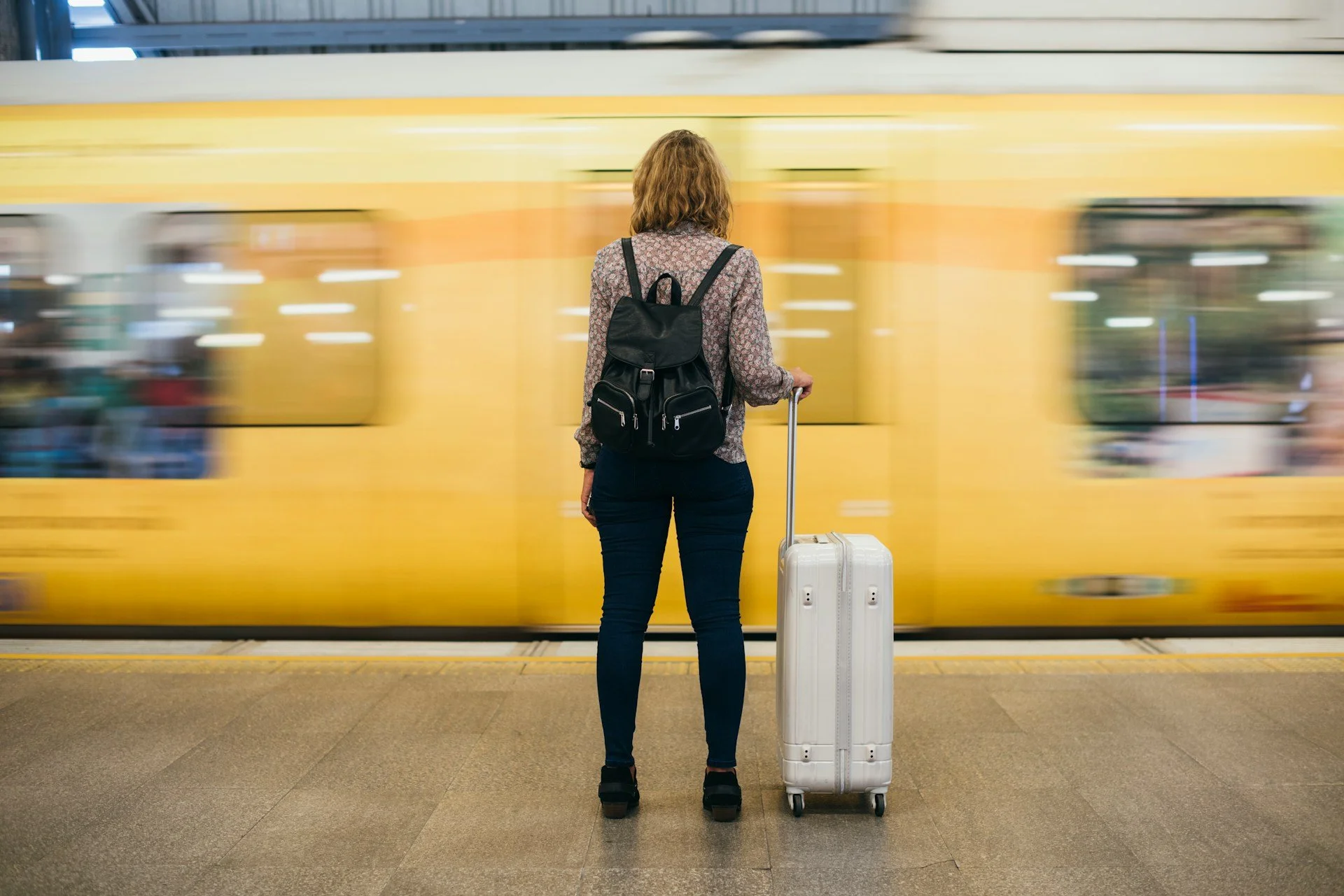A woman with shoulder-length hair, wearing a patterned shirt and dark pants, stands on a train platform facing a yellow train. She has a black backpack and is holding a white rolling suitcase, waiting as the train speeds by.
