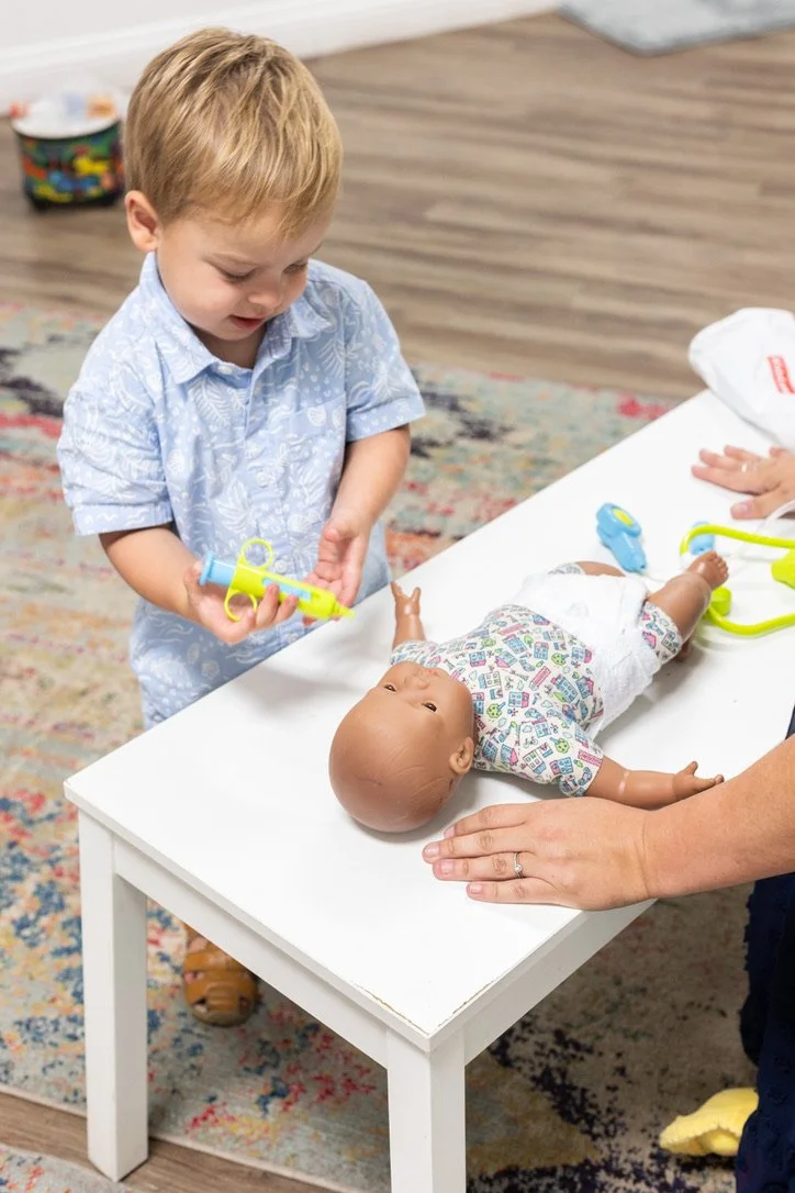 A young boy playing doctor with a baby doll on a white table. The boy is holding a toy syringe and wearing a light blue shirt. An adult's hand is visible, gently holding the doll down.