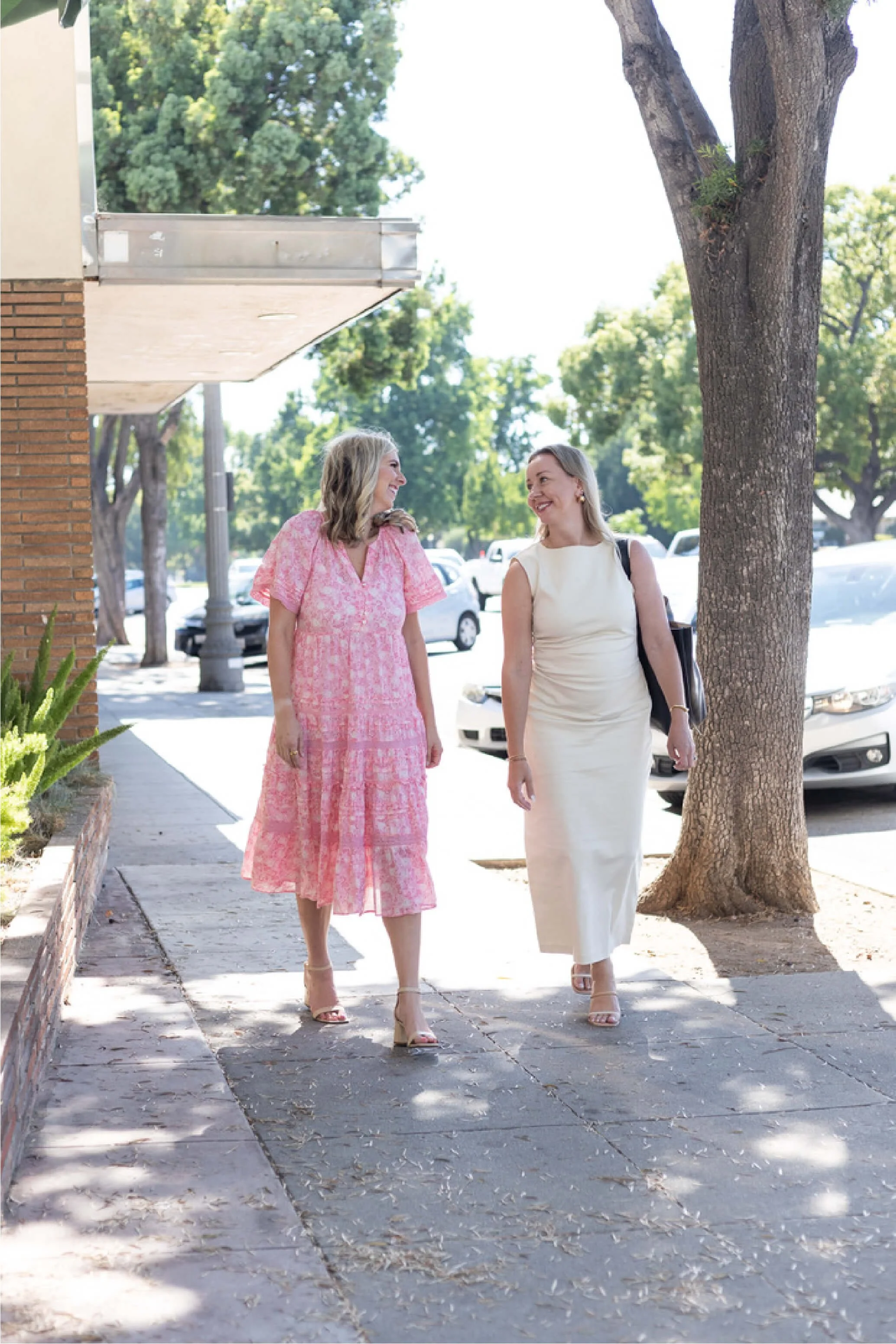 Two women walking on a sidewalk, one in a pink dress and the other in a white dress, smiling and talking to each other. There are trees, parked cars, and a brick building in the background.