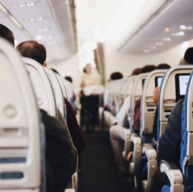 Inside an airplane cabin with passengers seated and a flight attendant standing at the front.