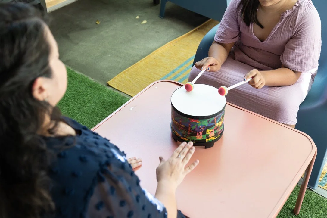 Two girls sitting at a pink table playing a musical toy with drumsticks.