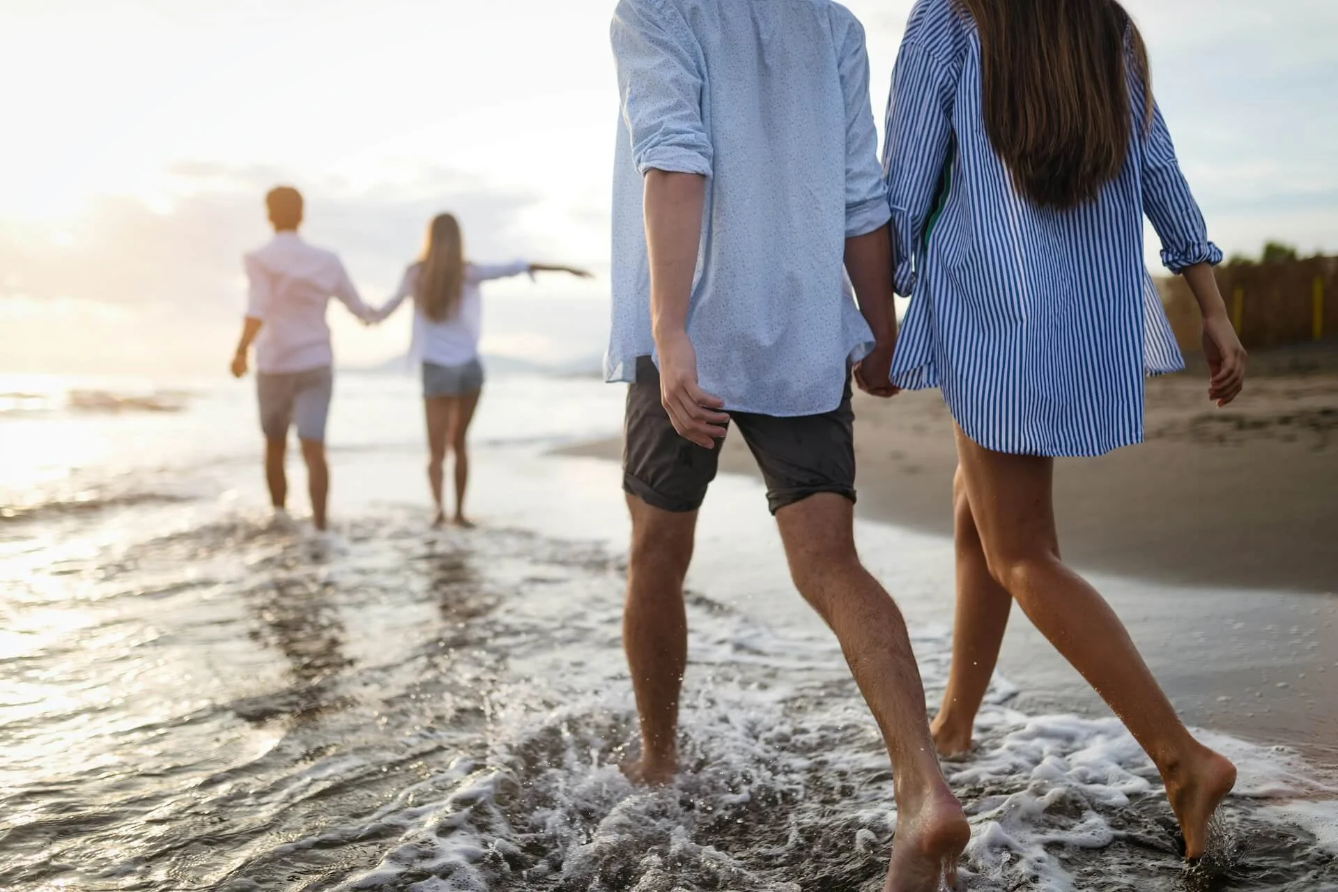 People walking along the shoreline at the beach during sunset, with two individuals holding hands in the foreground and two others in the background.