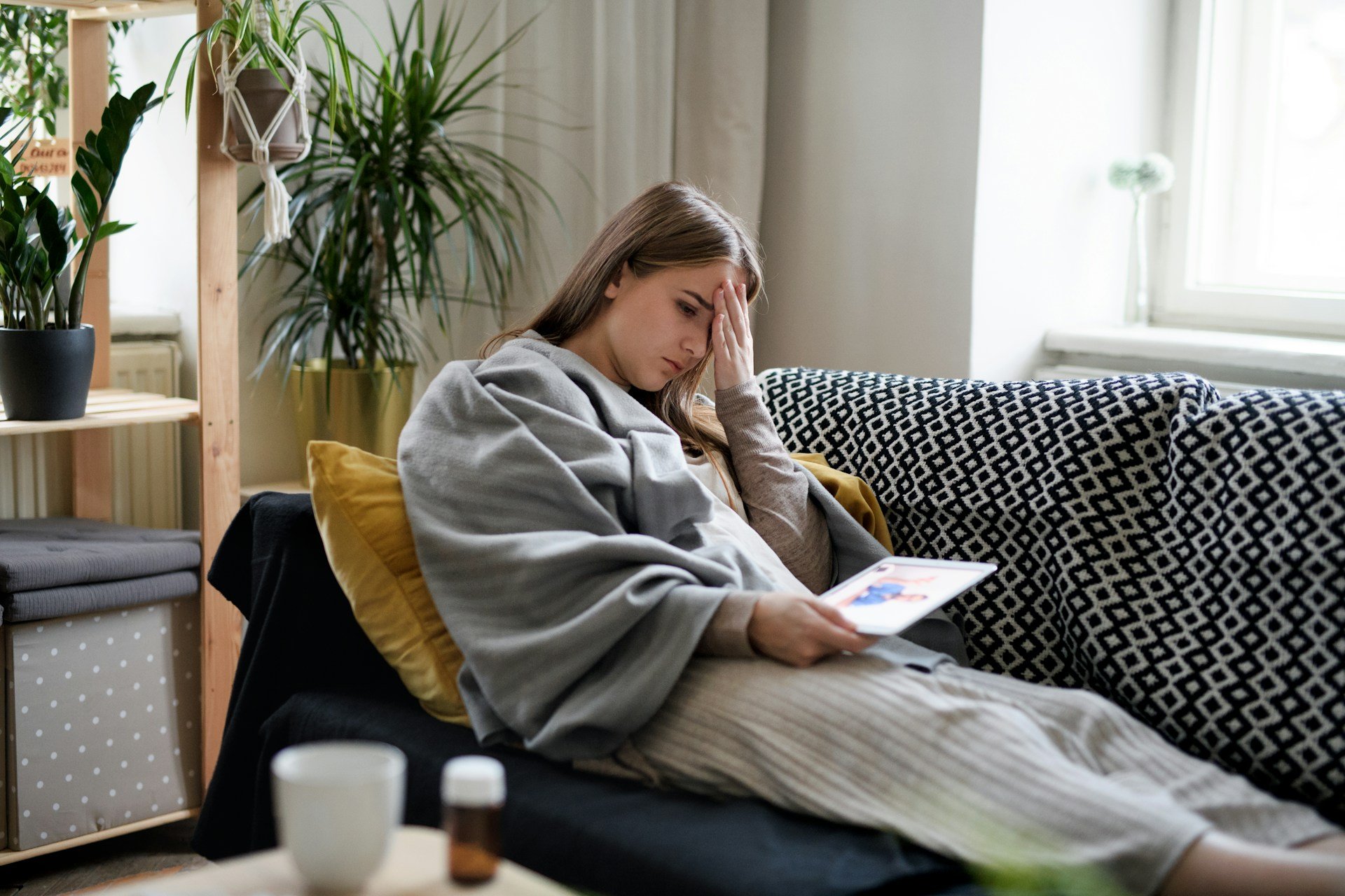 A woman sitting on a sofa in a cozy living room, holding a tablet and looking concerned, with plants and home decor in the background.
