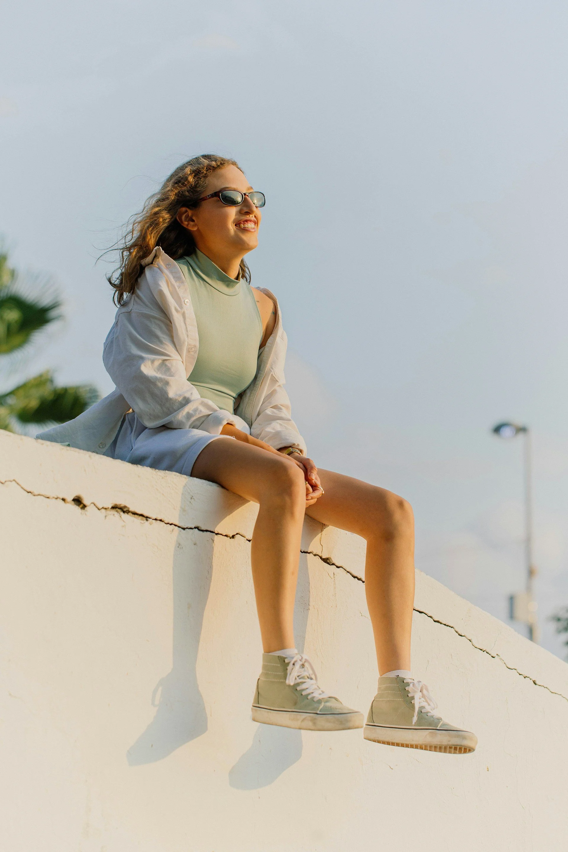 A woman sitting on a white wall, wearing sunglasses, a light green tank top, a cream jacket, and beige high-top sneakers, smiling and enjoying the sunny weather.