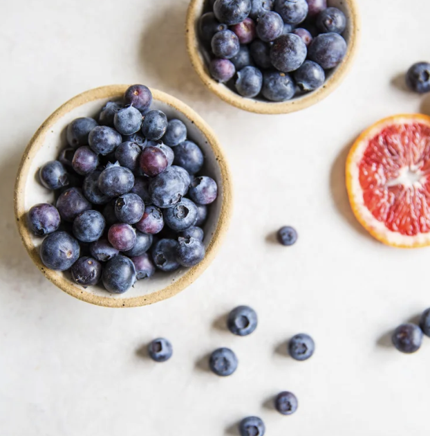 Two small bowls filled with blueberries and a sliced half of a blood orange on a white surface with loose blueberries scattered around.