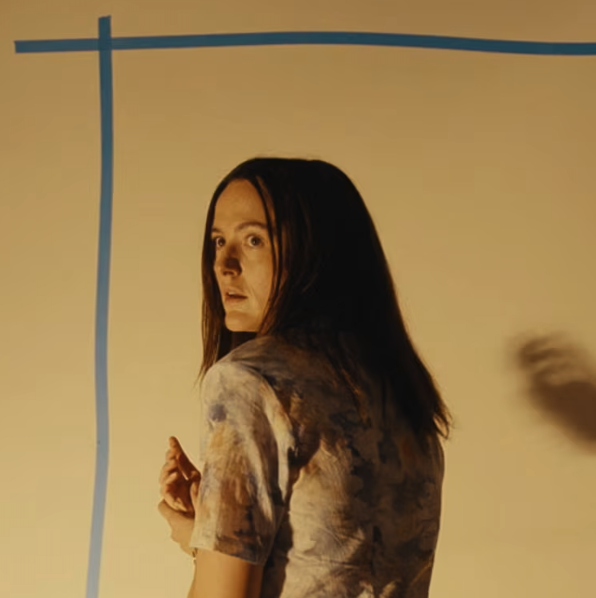 A woman with long dark hair looking over her shoulder against a beige background with blue lines.
