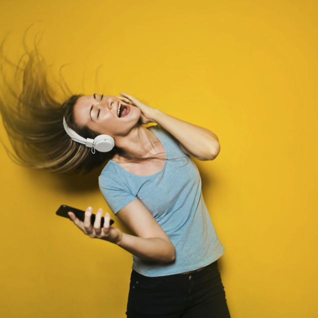 A woman with long hair wearing a gray t-shirt and headphones, holding a phone, dancing and enjoying music against a yellow background.