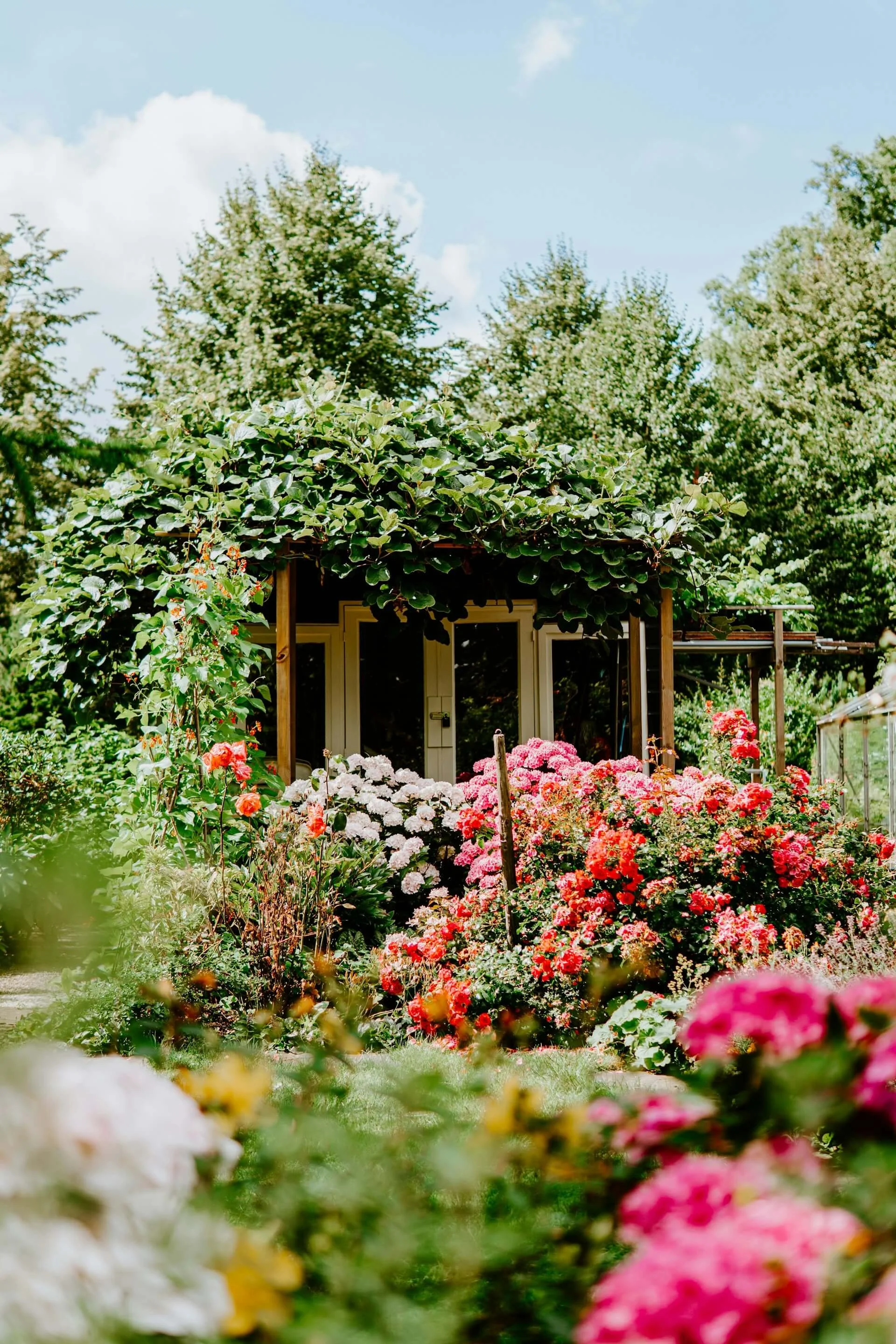 A lush garden with colorful flowers in the foreground, a small house with large windows and a door surrounded by greenery, and tall leafy trees under a partly cloudy sky.