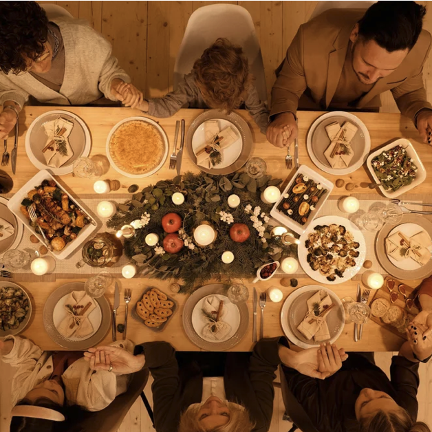 People sitting around a wooden dining table, children and adults, with a festive centerpiece of candles, pine branches, and red ornaments, set for a holiday meal with various dishes and desserts.