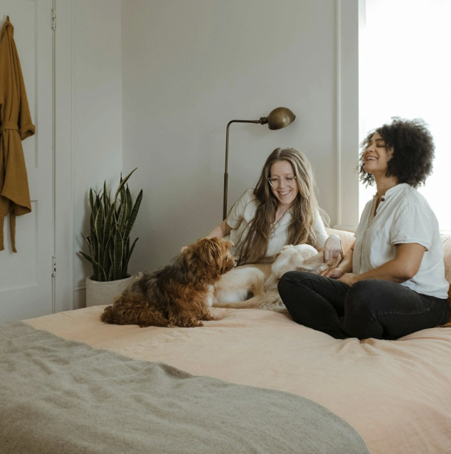 Two women sitting on a bed with their dogs, smiling and enjoying their time together in a cozy bedroom.
