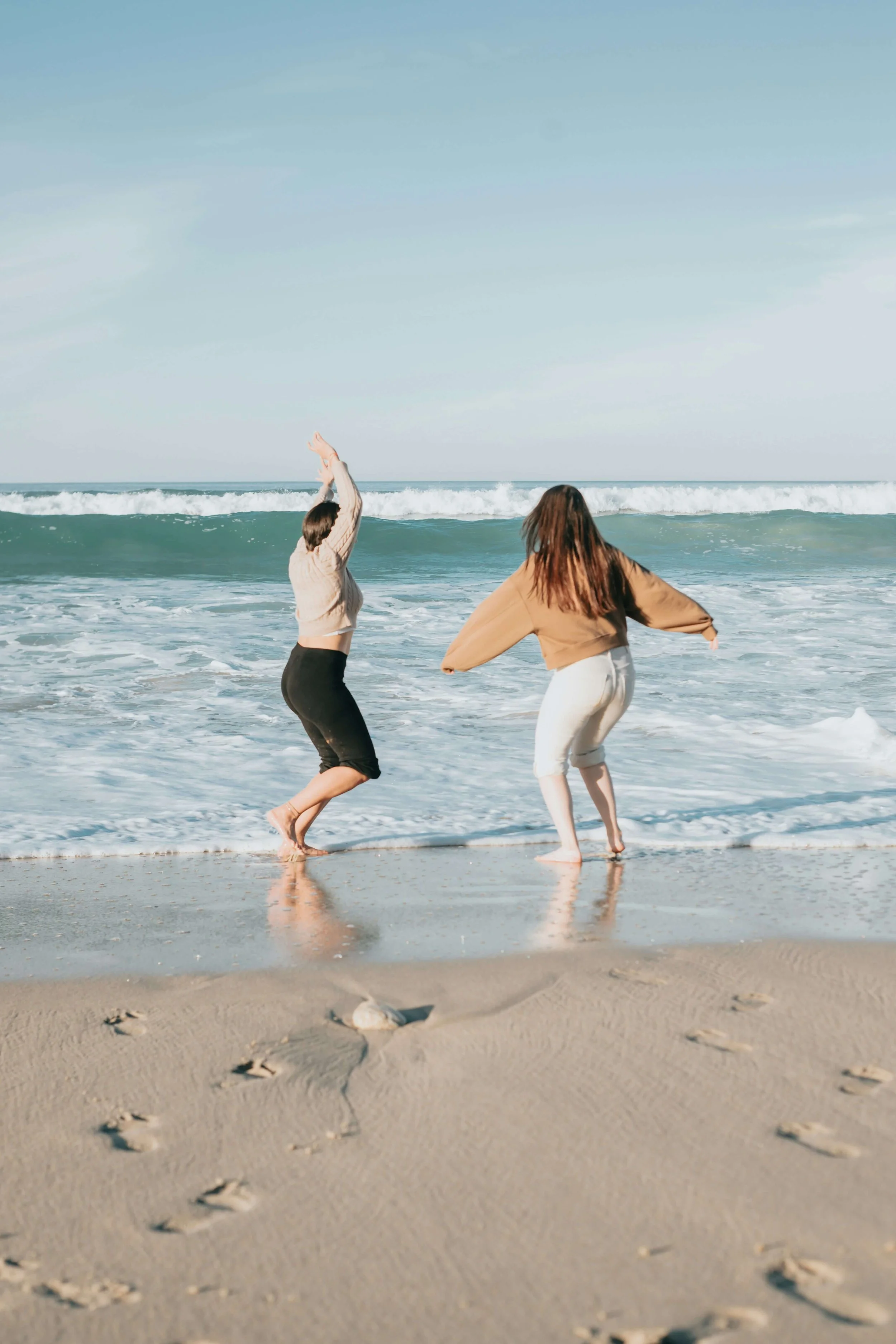 Two women stand on a sandy beach near the ocean, facing away from the camera. One woman is raising her arms, and the other is bending slightly. The sky is clear with some light clouds, and there are footprints in the sand.