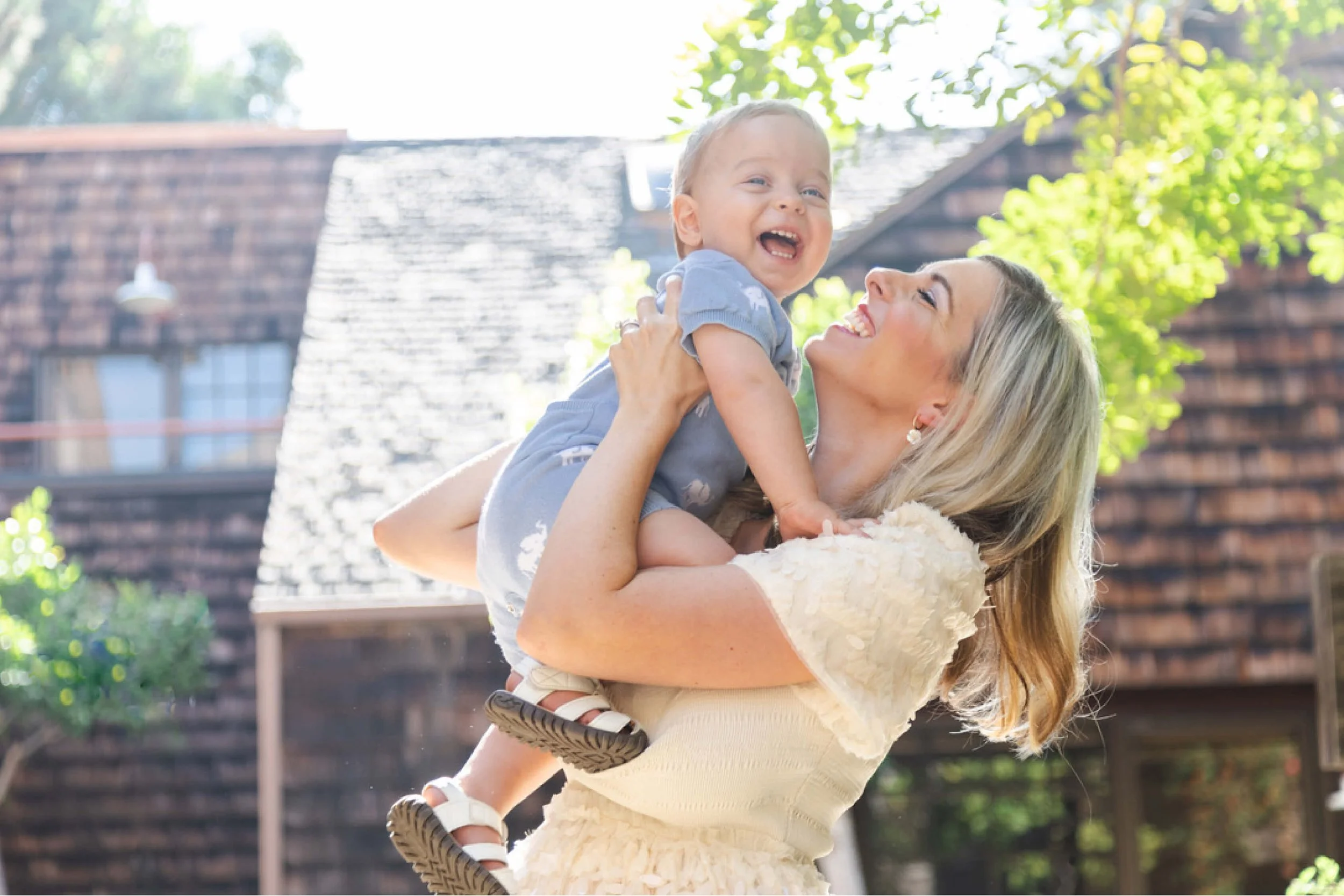 A woman with long blonde hair, wearing a beige dress, holding a young boy with short blonde hair, wearing a light blue shirt with white patterns, smiling and looking at the camera. They are outside in a backyard with a brick house, trees, and sunlight.