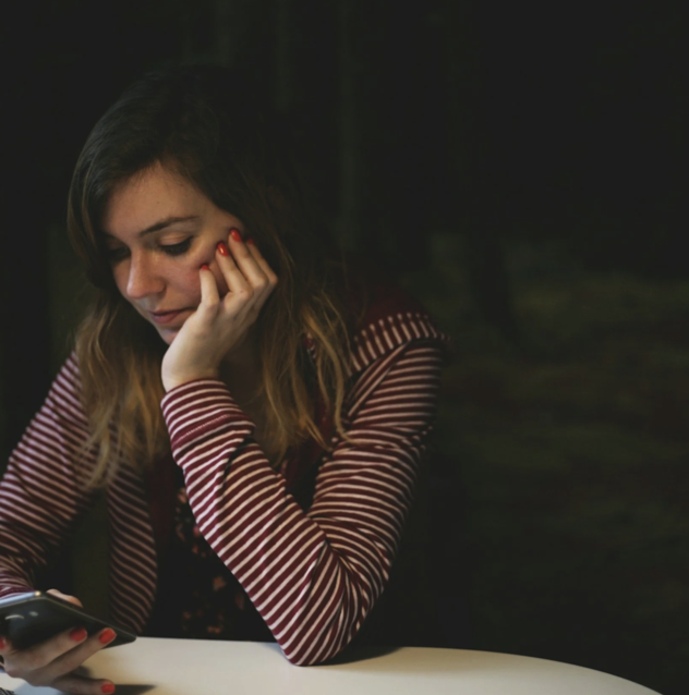 A young woman with long, wavy hair sitting at a table, looking at her phone with a thoughtful expression in a dark environment.