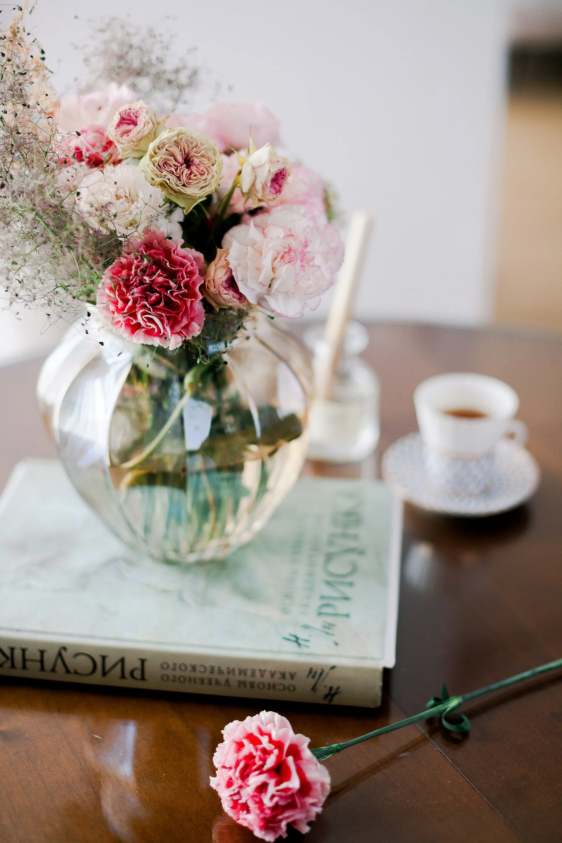 A glass vase with a bouquet of pink and white flowers, placed on top of a book on a wooden table. There is a cup of tea or coffee and a pink carnation flower lying on the table.