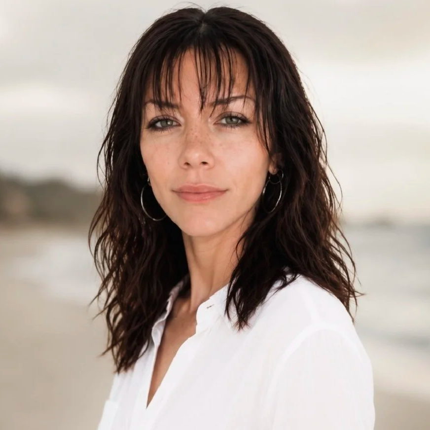 Close-up of a woman with dark wavy hair, wearing hoop earrings and a white shirt, at the beach with blurred shoreline in the background.