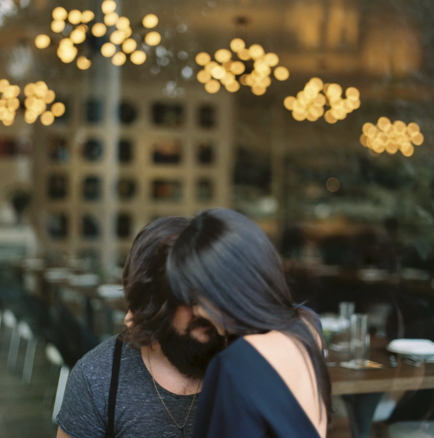 A couple sitting close together at a table in a restaurant or cafe, sharing a tender moment with foreheads touching. The background features warm string lights and blurred interior decor.