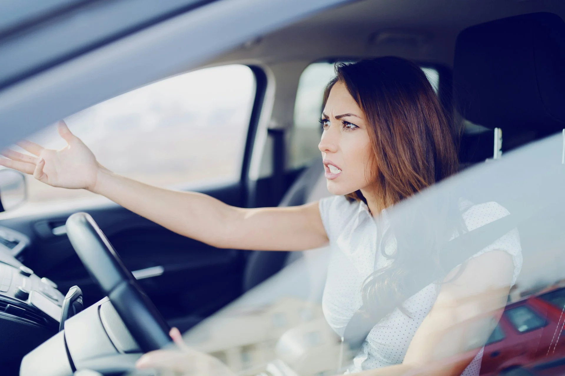 Angry woman driving a car, reaching toward the window with a frustrated expression.
