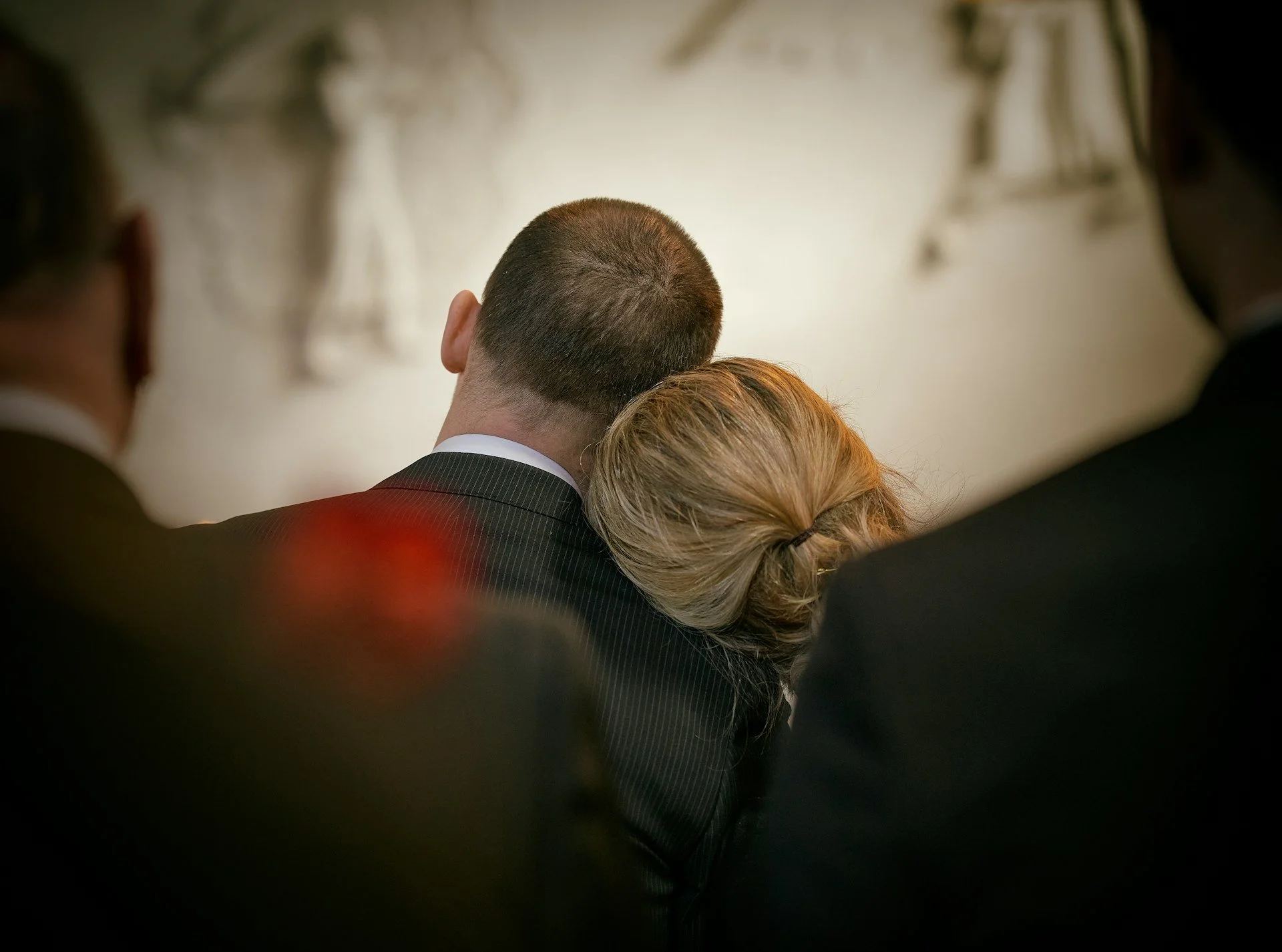 Back of a person's head with short hair, surrounded by blurred figures in a formal setting.